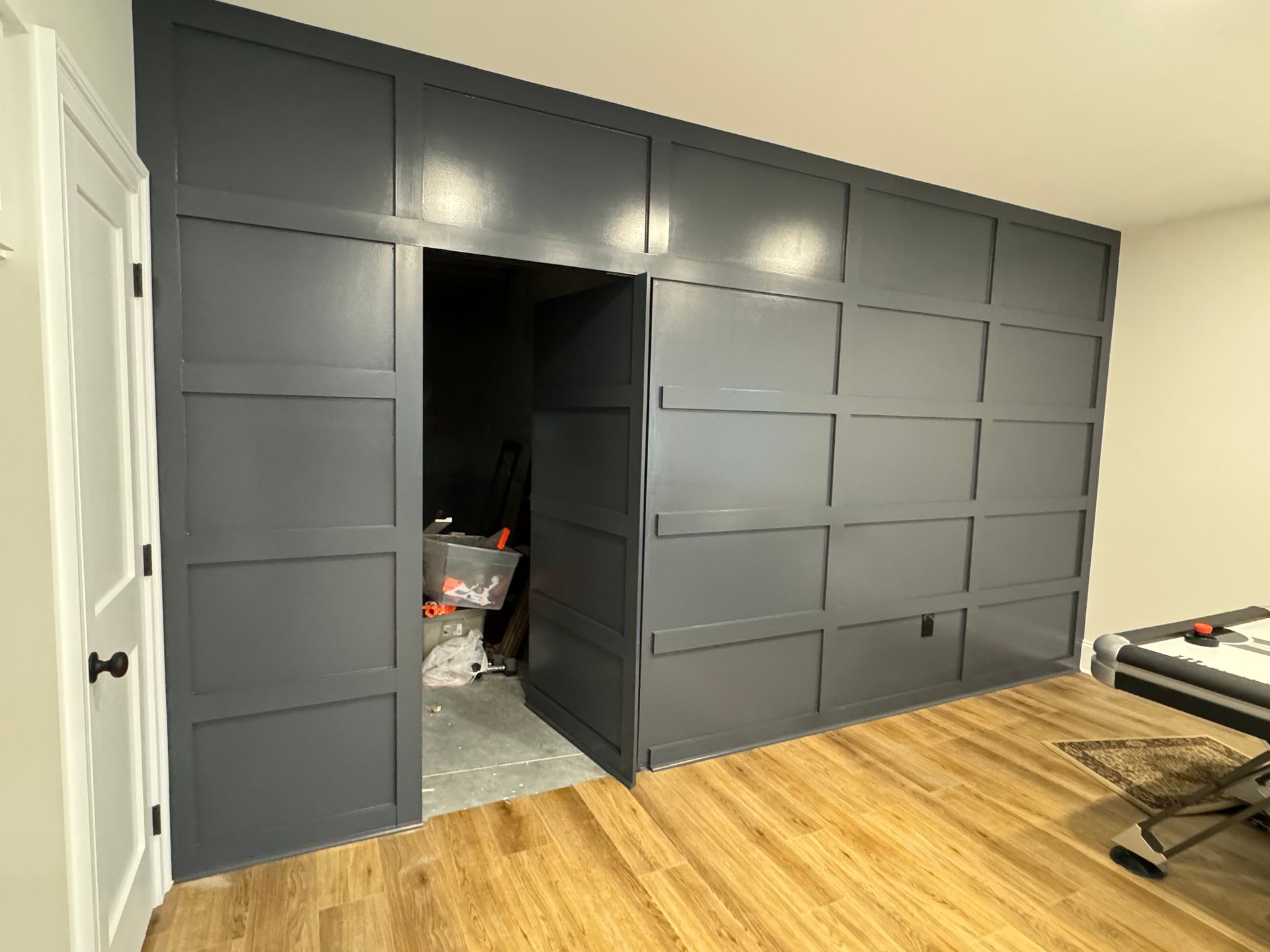 A dark grey grid-paneled accent wall with a hidden, door-sized opening leading into a storage space in a wooden floor room.