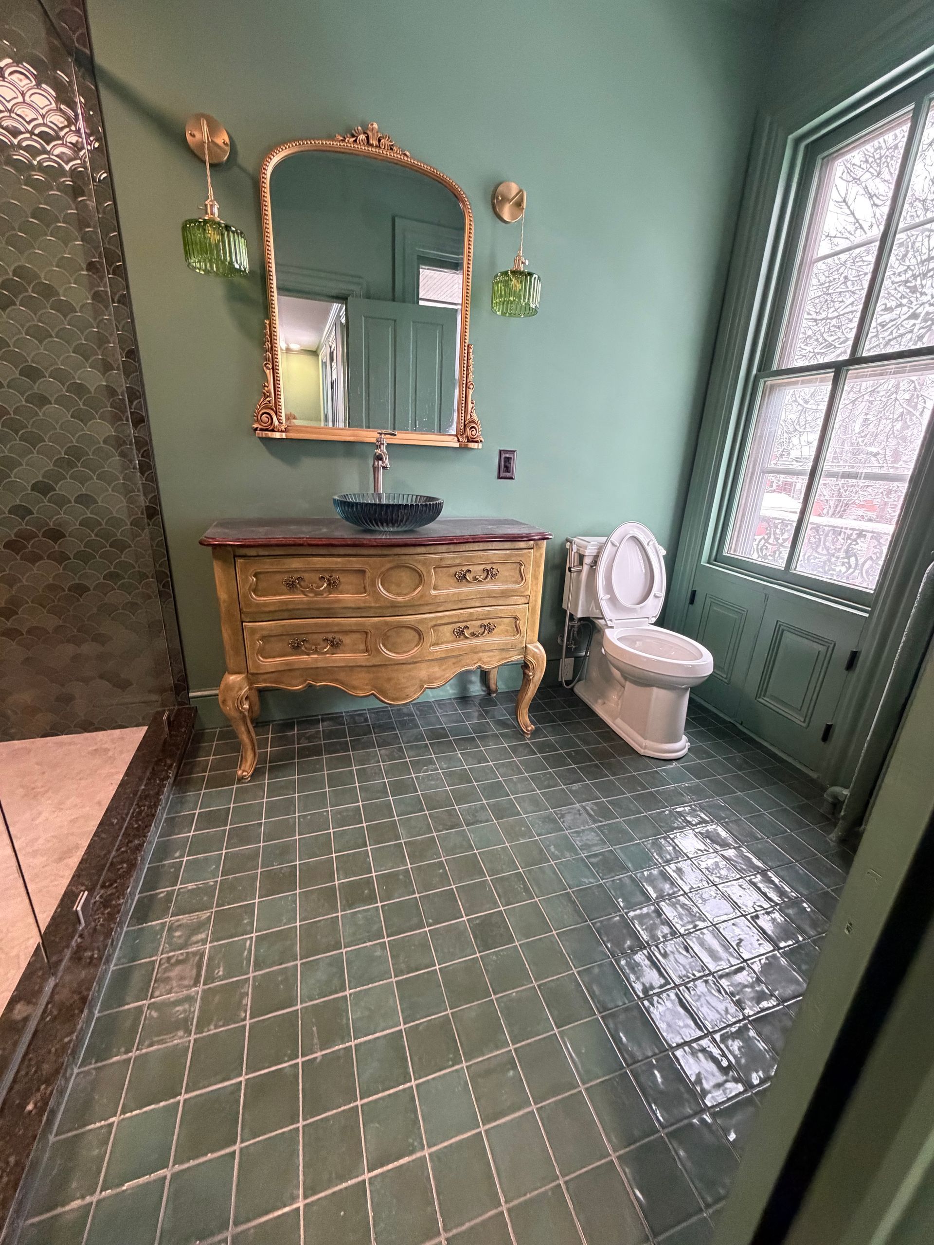 A green-walled bathroom featuring a gold antique vanity with a dark basin, a matching mirror, and a window with trim.