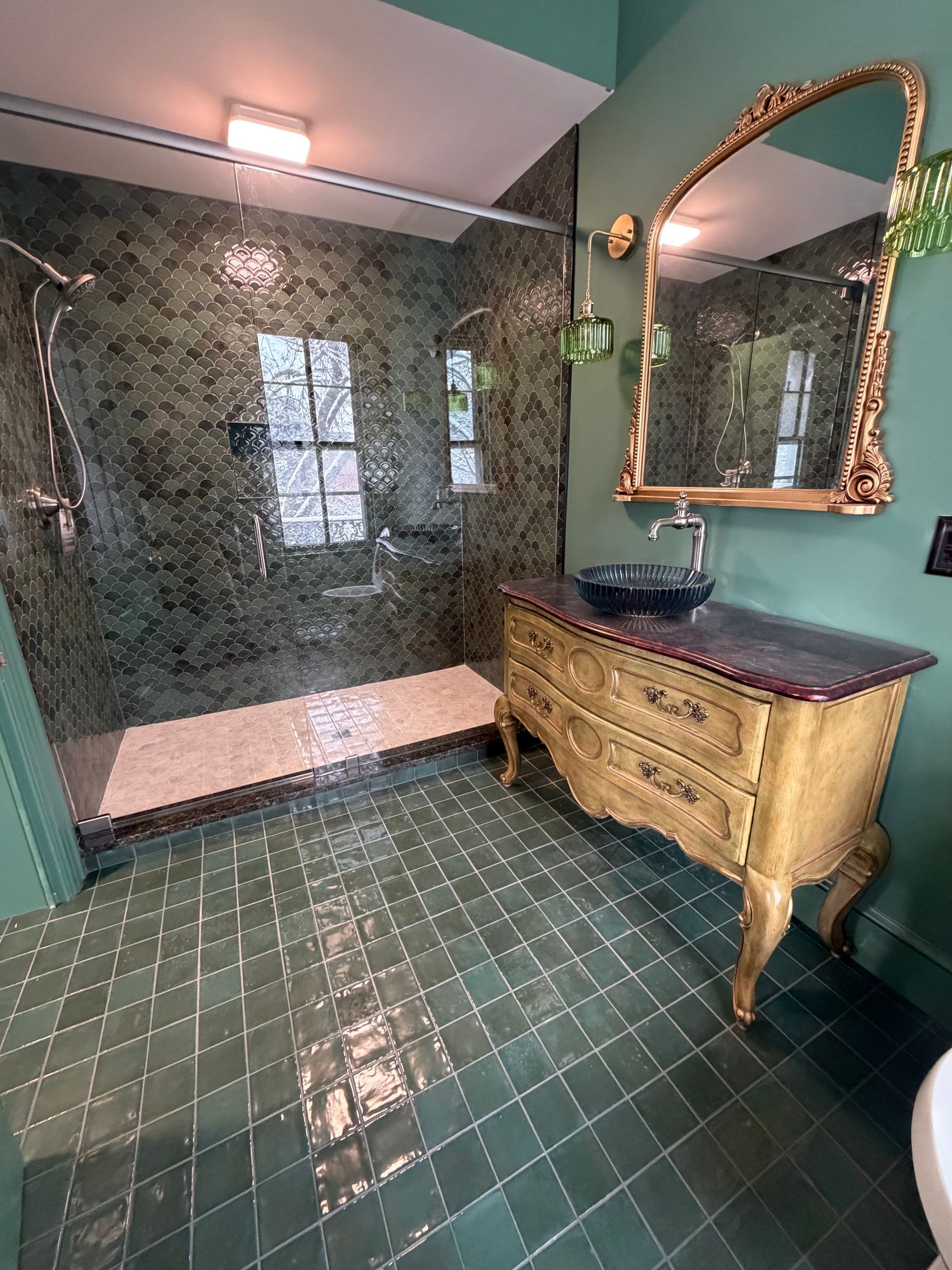 A luxurious bathroom with green-tiled floors, a walk-in shower with dark patterned walls, and a vintage wooden vanity.