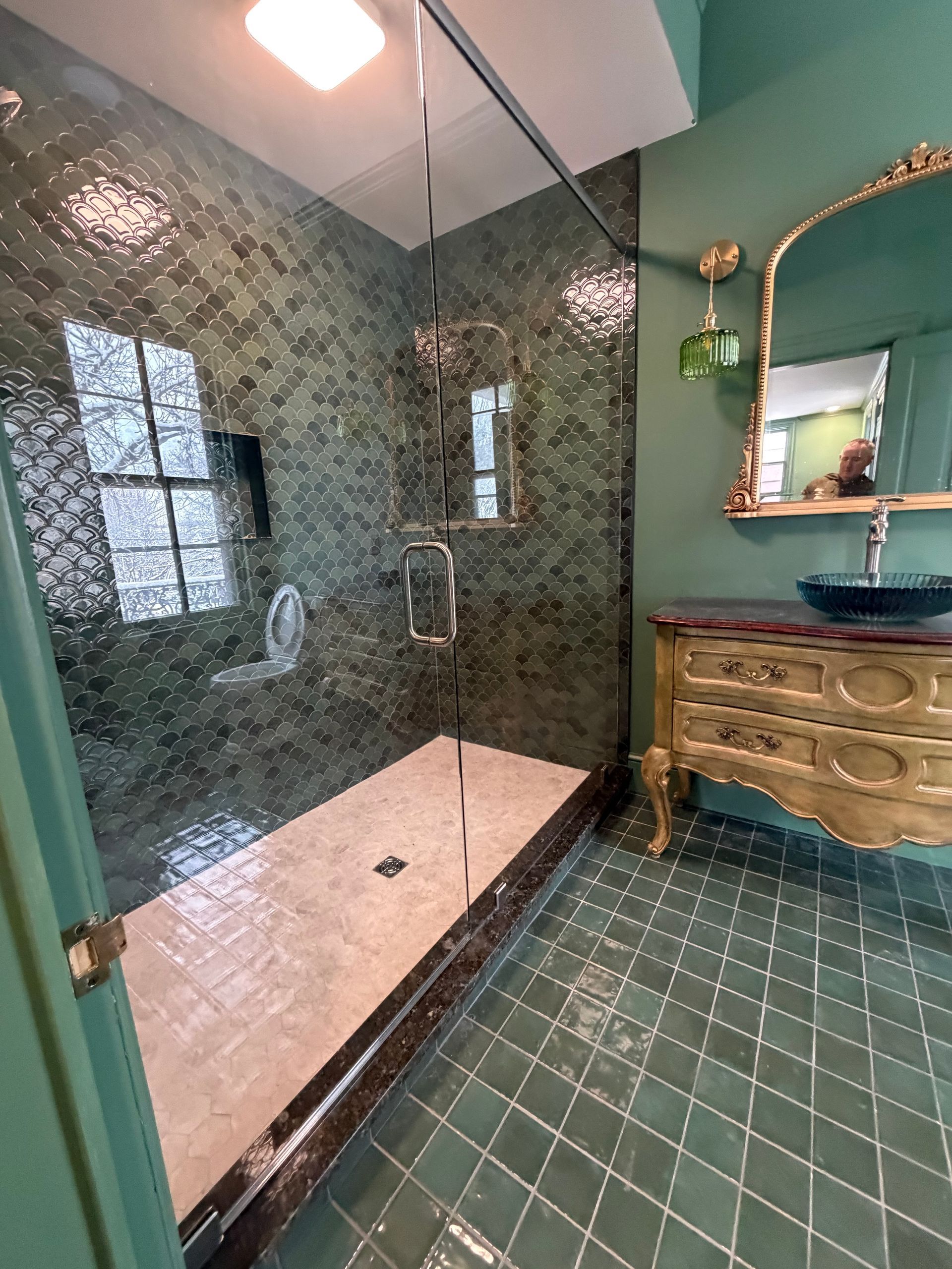 A bathroom with dark green wall and floor tiles, a walk-in glass shower, and an ornate gold vanity with a mirror.