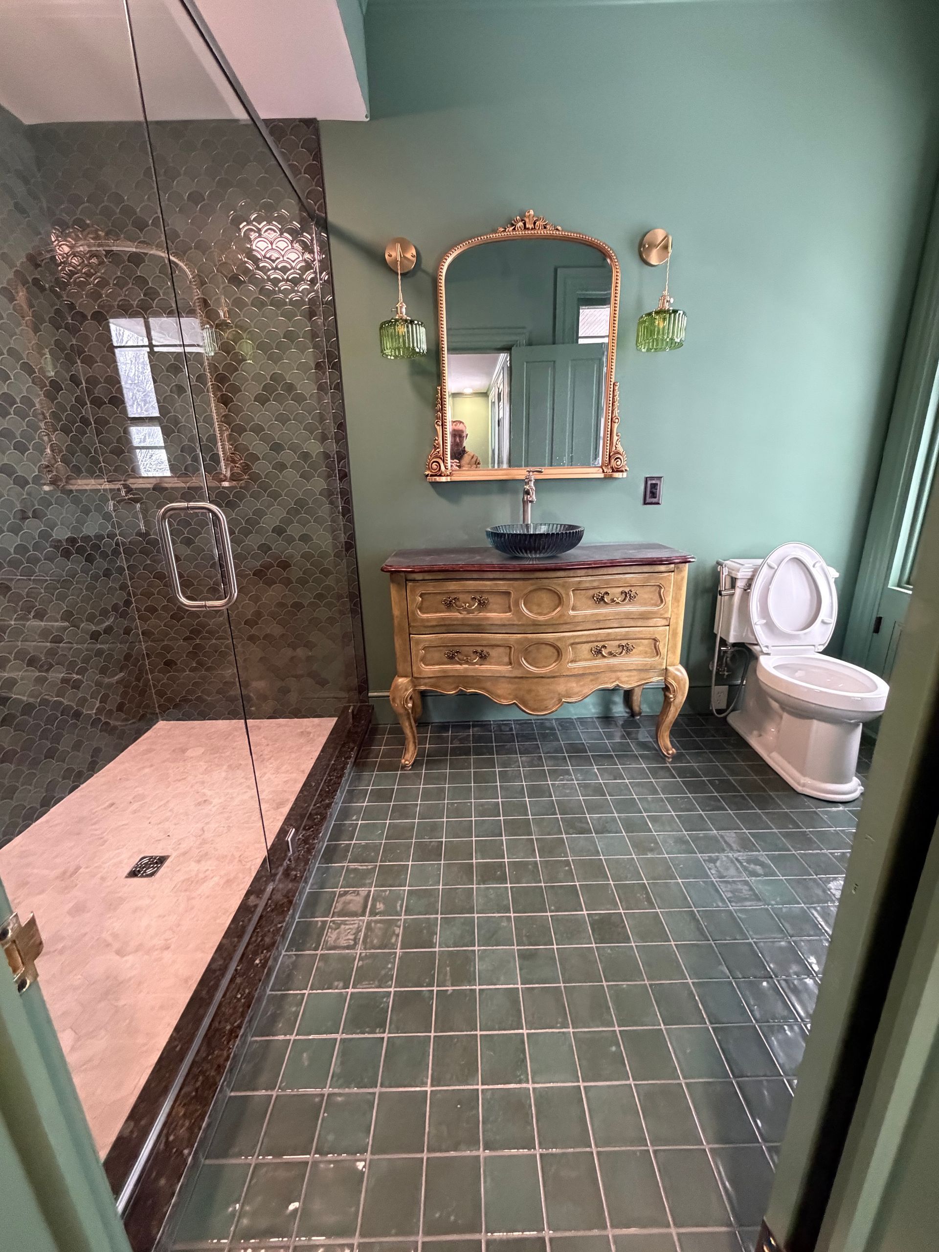A green-walled bathroom with a dark tiled shower, a vintage wooden vanity with a vessel sink, and a white toilet.