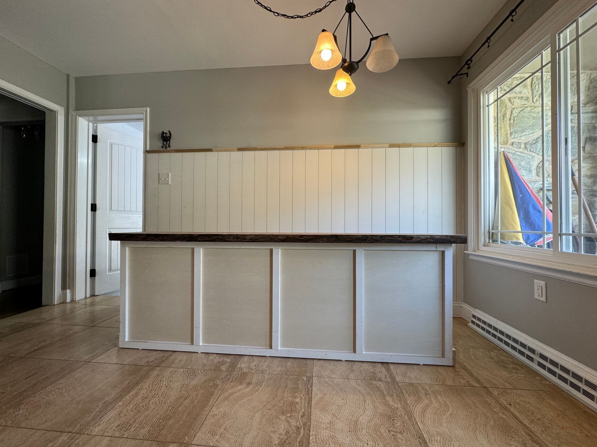 A view of a home bar with a white base, wood countertop, and shiplap wall, under a three-light fixture in a room.