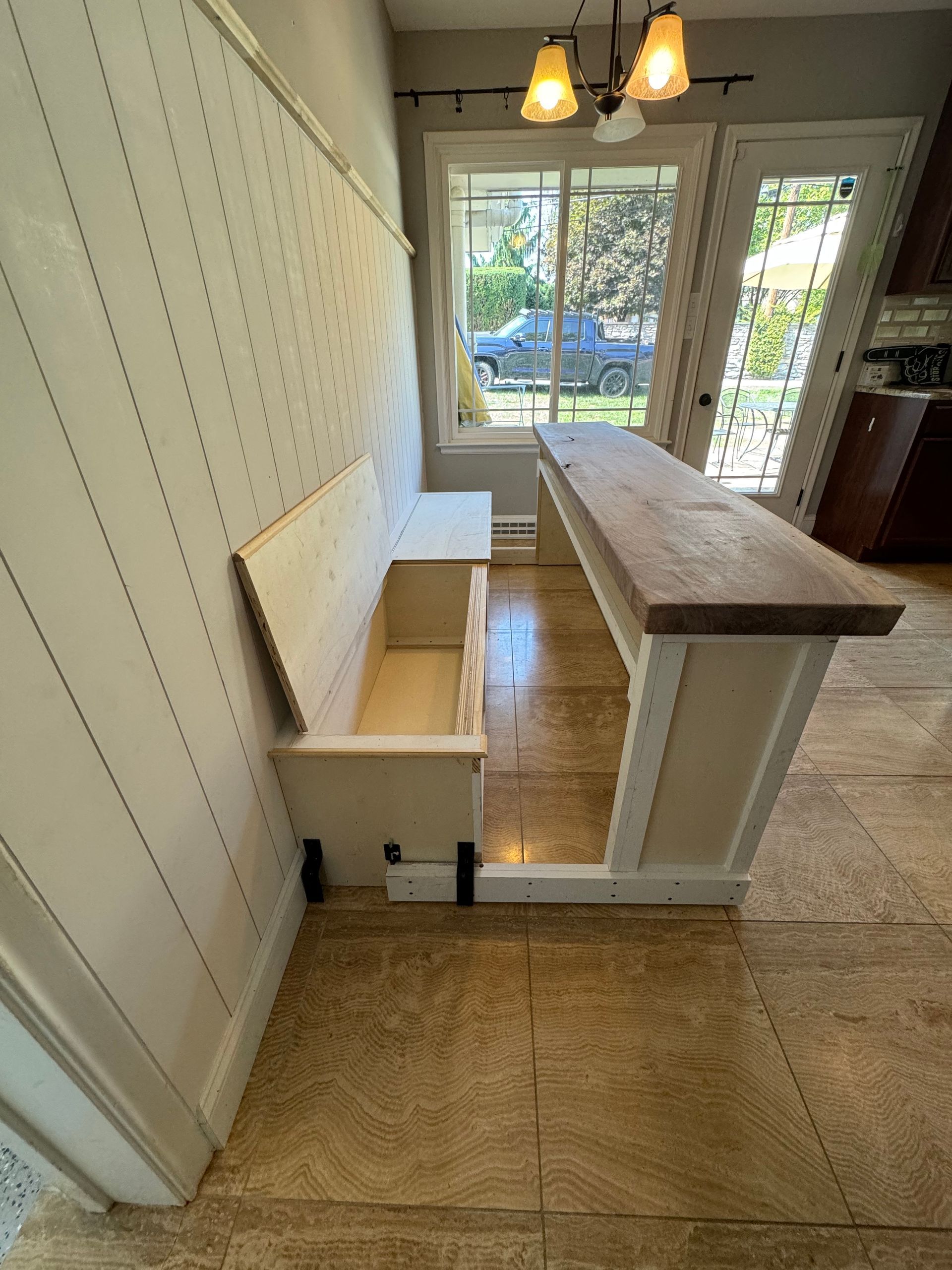 A white storage bench with an open lid sits next to a wooden breakfast bar in a kitchen area with light flooring.