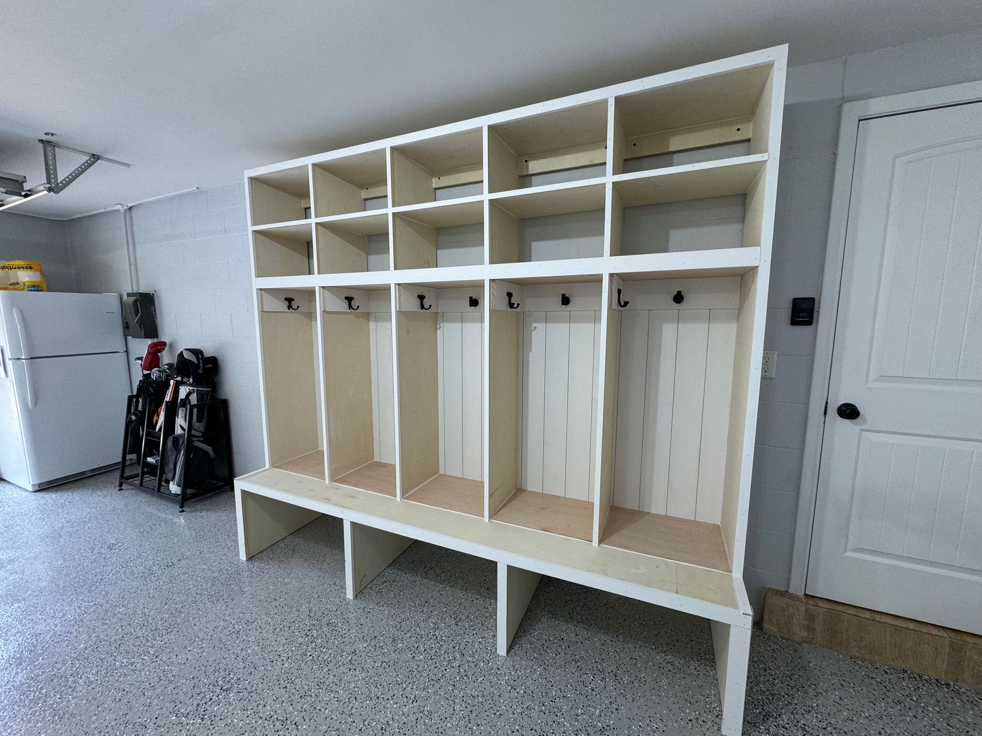 A white wooden locker unit with cubbies and coat hooks installed in a garage with a speckled floor.