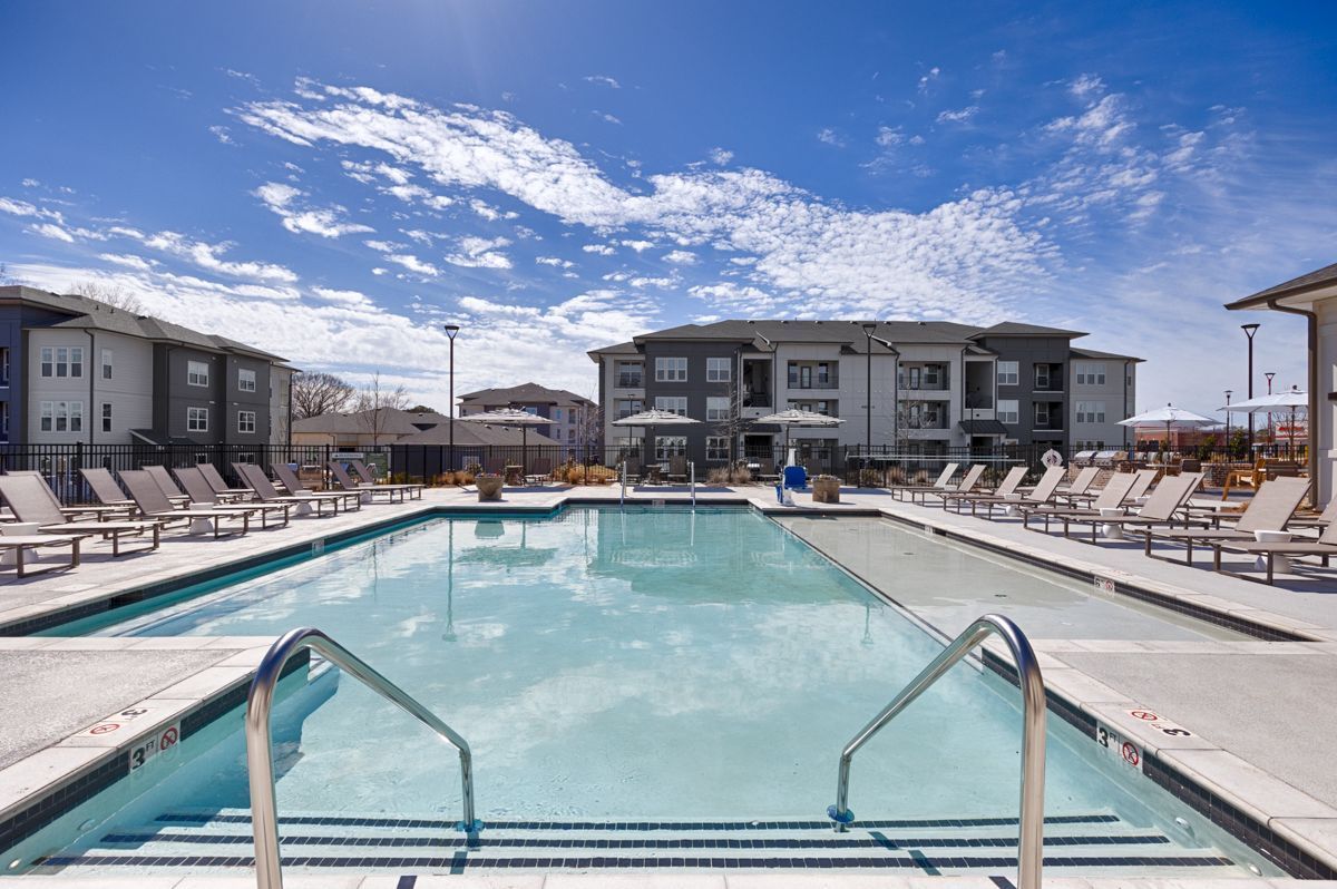 Pool in front of multi-story apartment buildings under a bright blue sky with lounge chairs.
