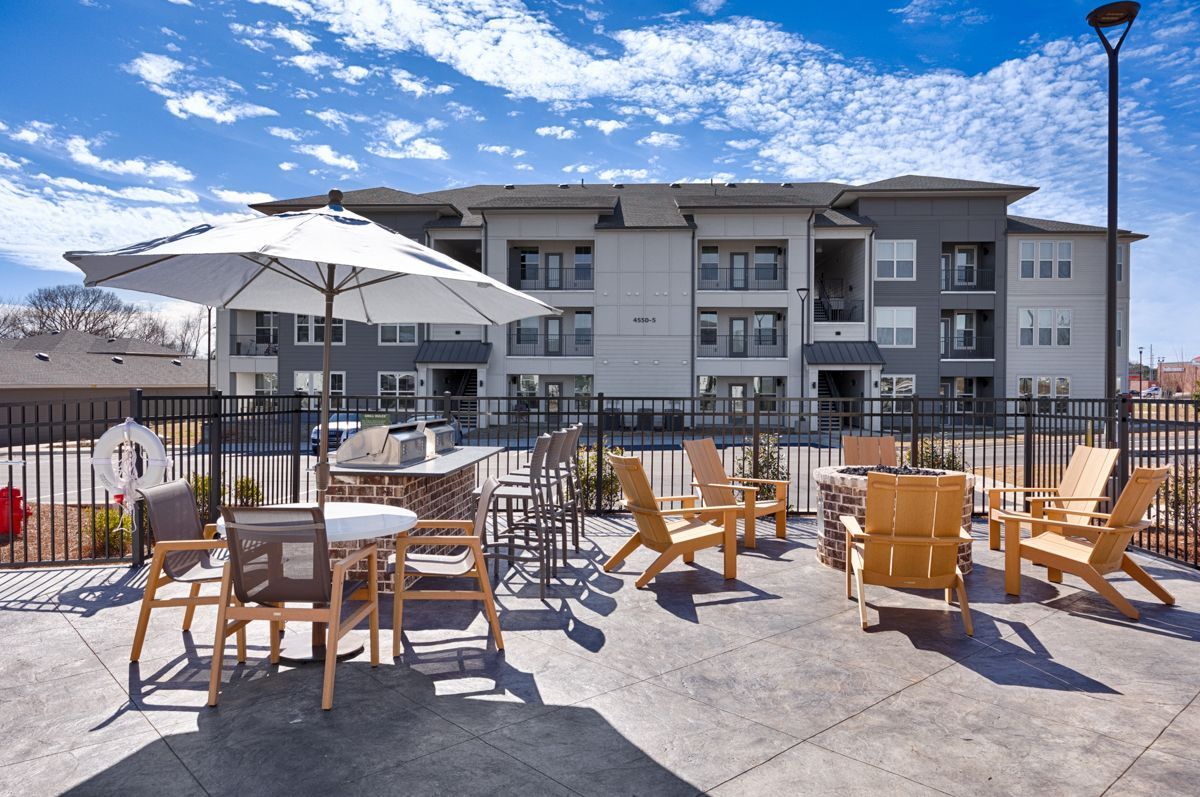 Outdoor patio with seating, grill, umbrella, and apartment building in the background. Sunny day.