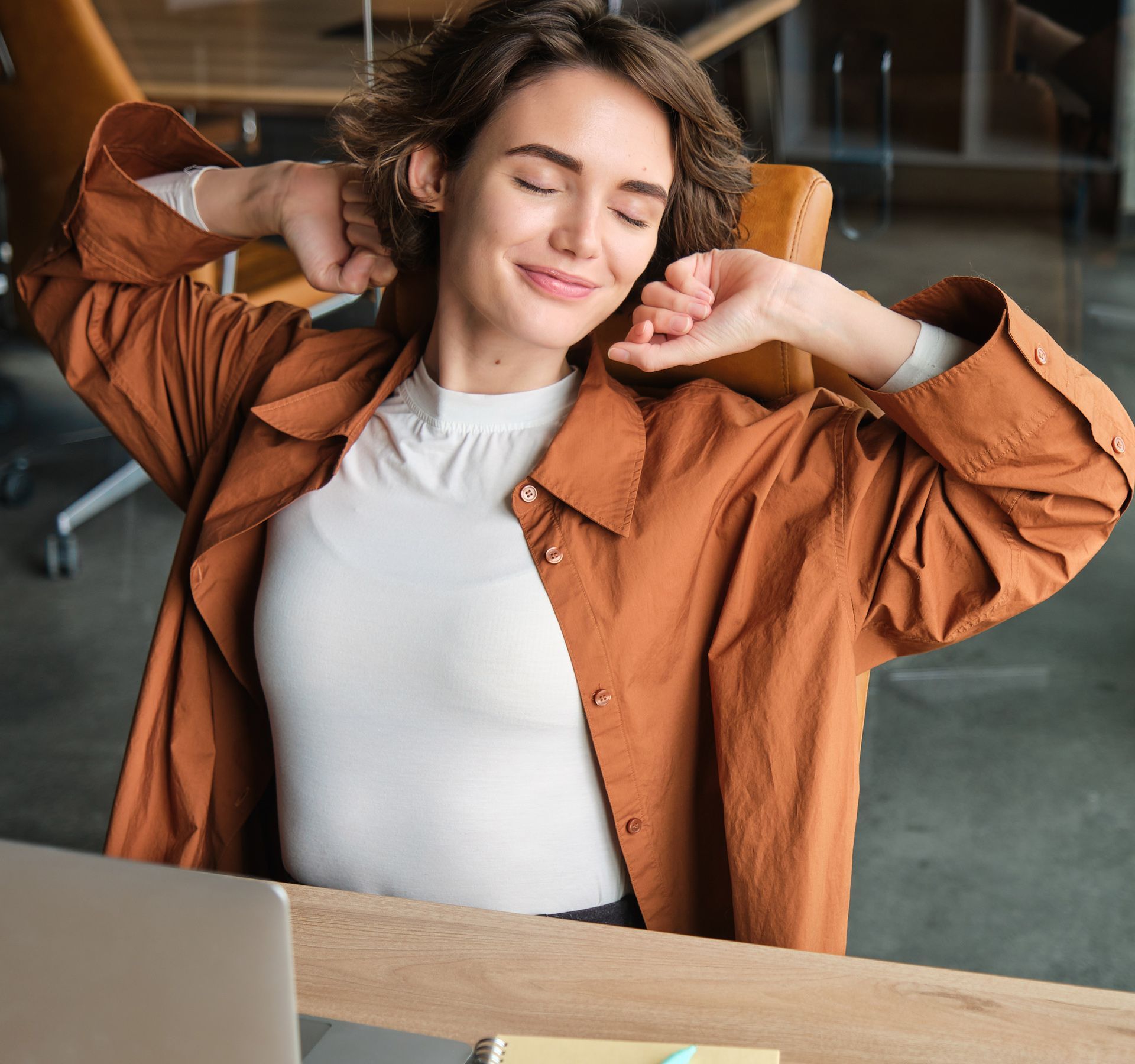Smiling woman stretching in an office, wearing a rust jacket and white shirt, seated at a desk with laptop