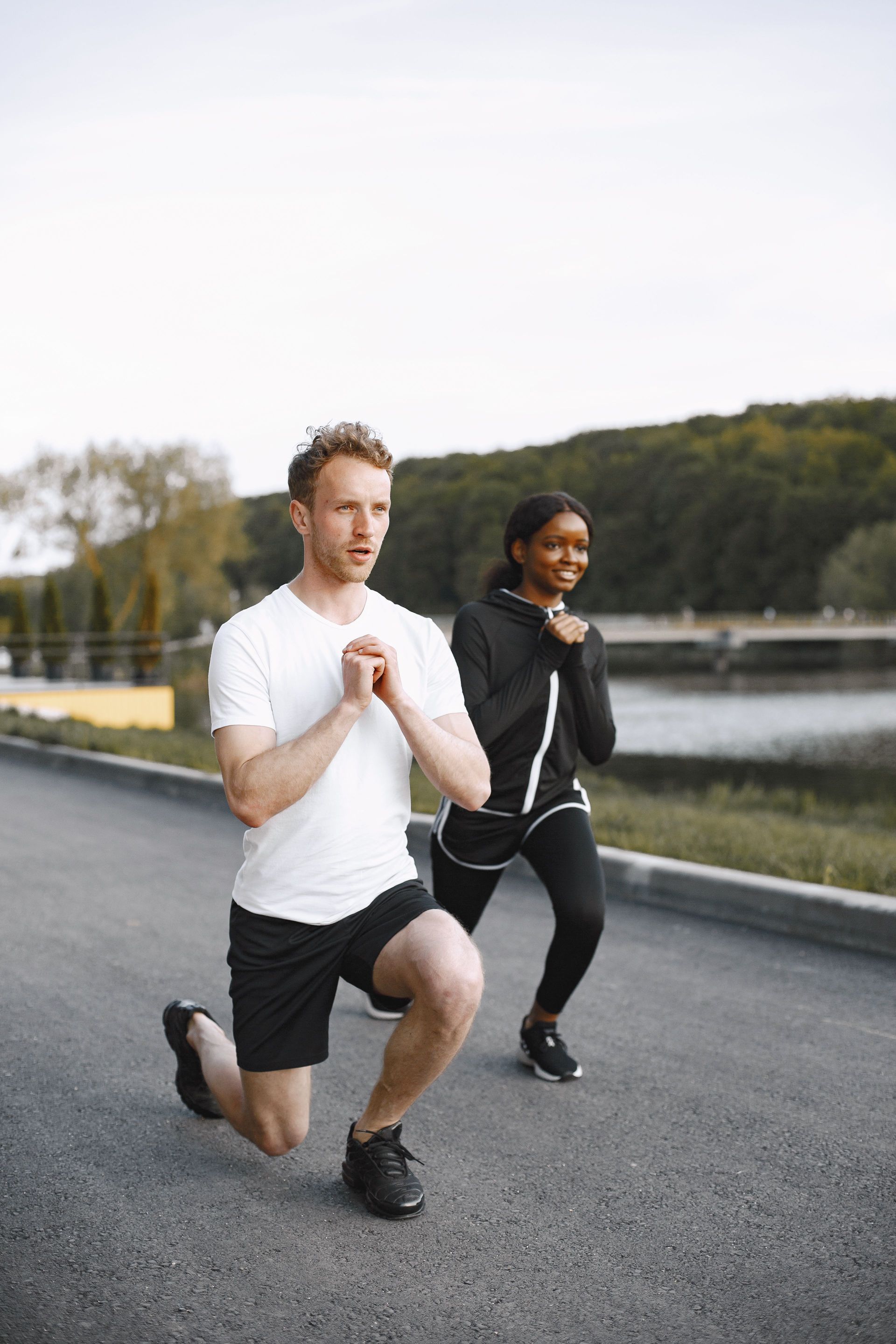 Two people doing walking lunges on a road beside water and trees