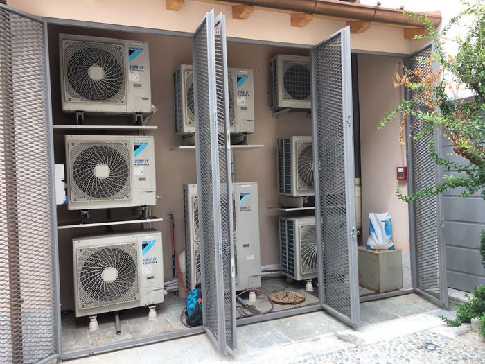 Unità compressore condizionatore esterne montate su una parete dell'edificio, con due cancelli metallici aperti sul davanti.