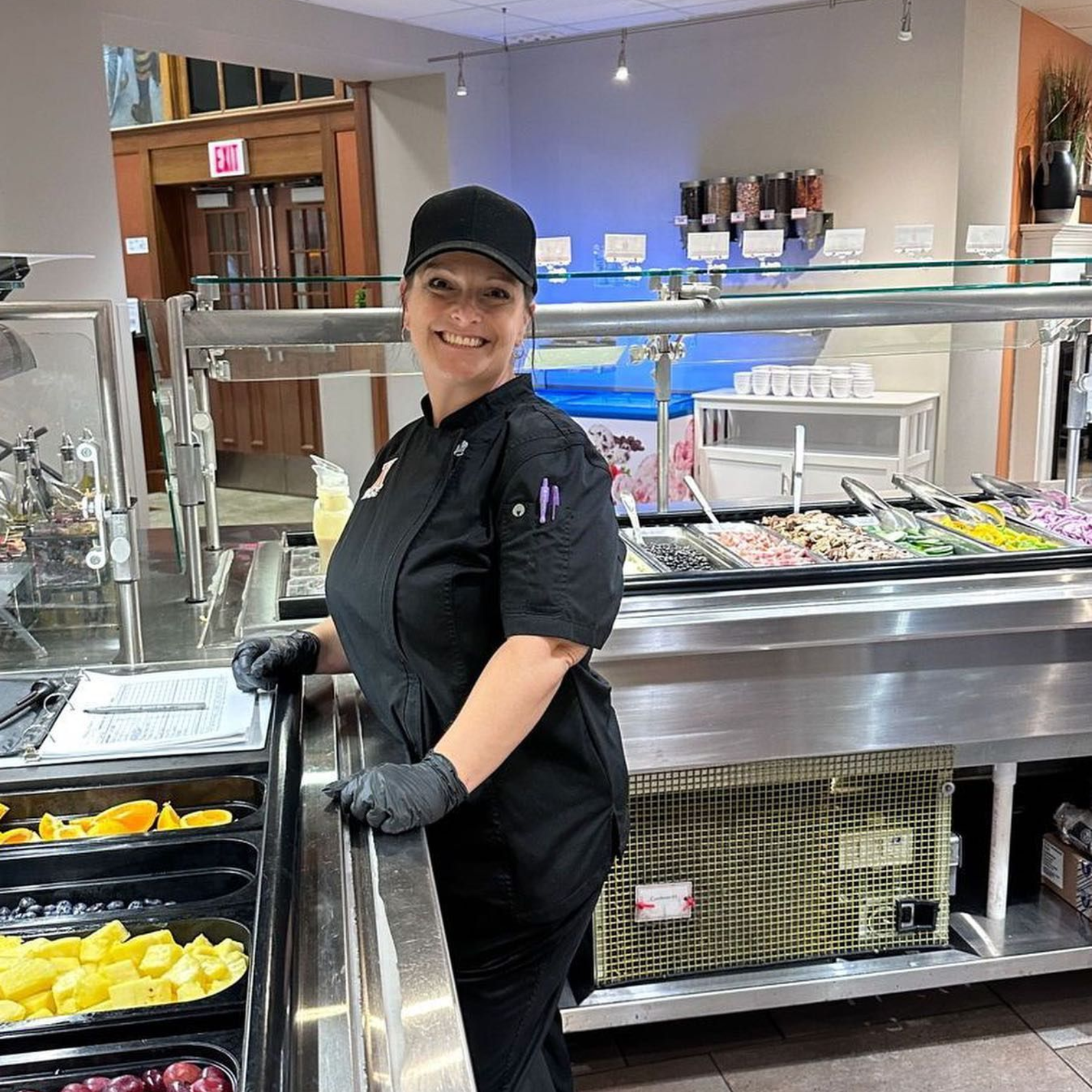 Chef in black uniform smiles next to a salad bar with various toppings and containers of fruit.