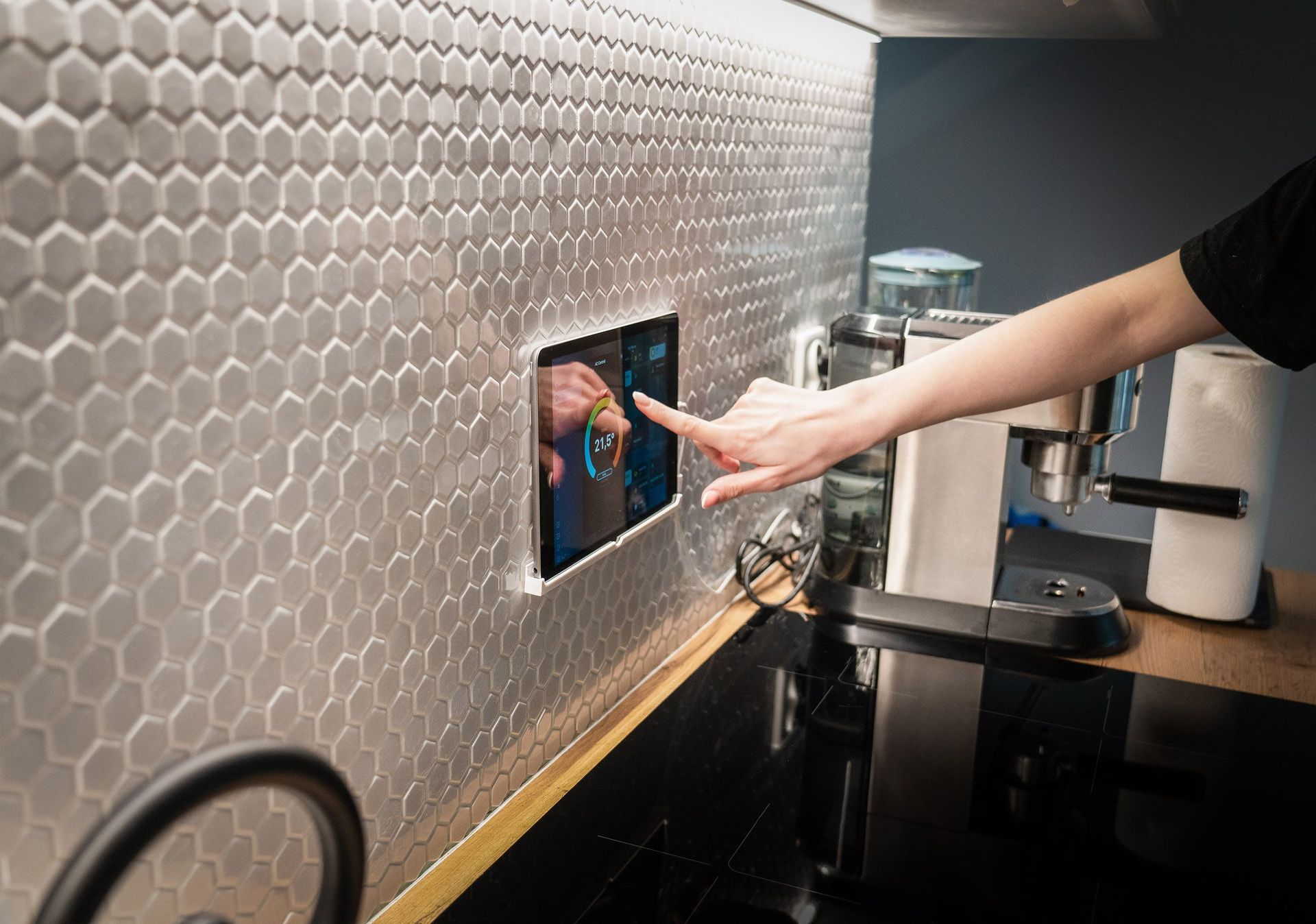Una persona allunga la mano verso uno schermo touchscreen montato a parete in una cucina moderna.
