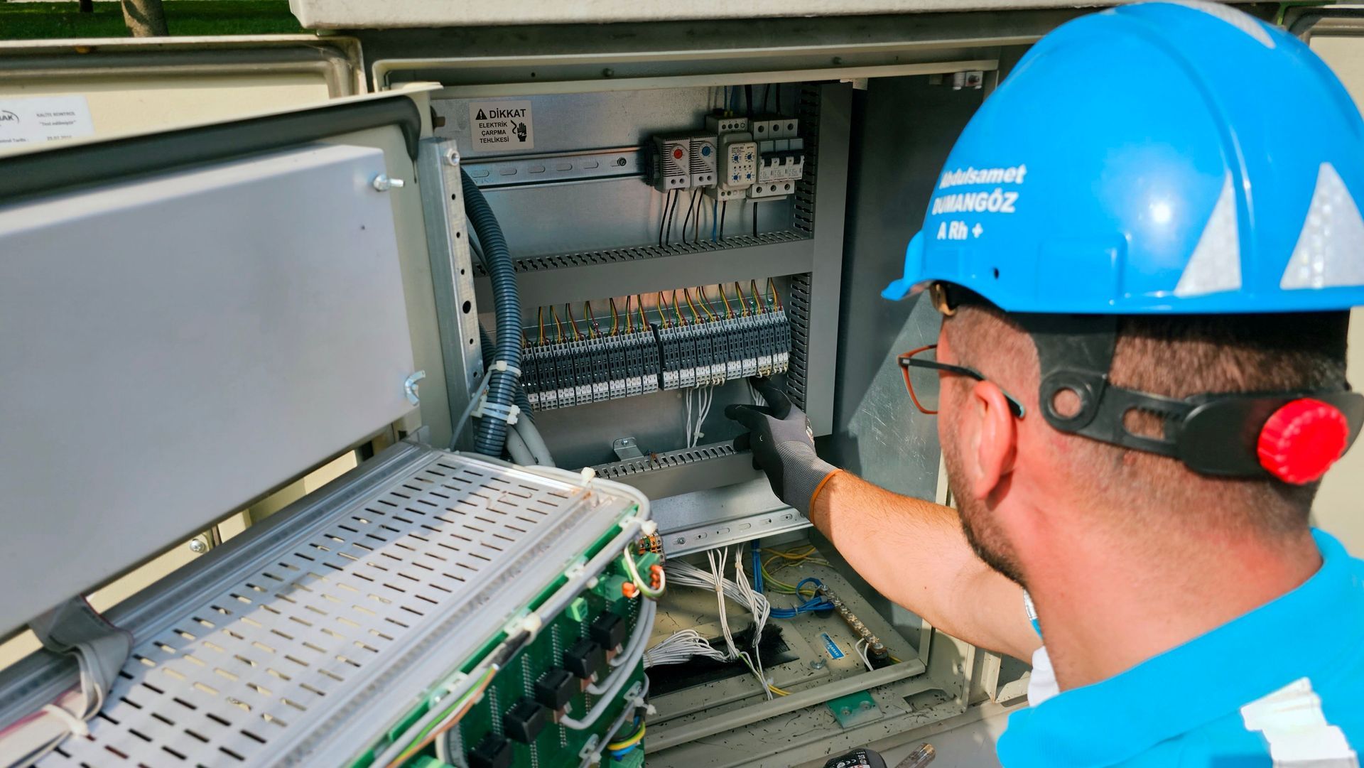Tecnico con elmetto blu ispeziona un quadro elettrico di controllo con cablaggi e pannelli di circuiti.