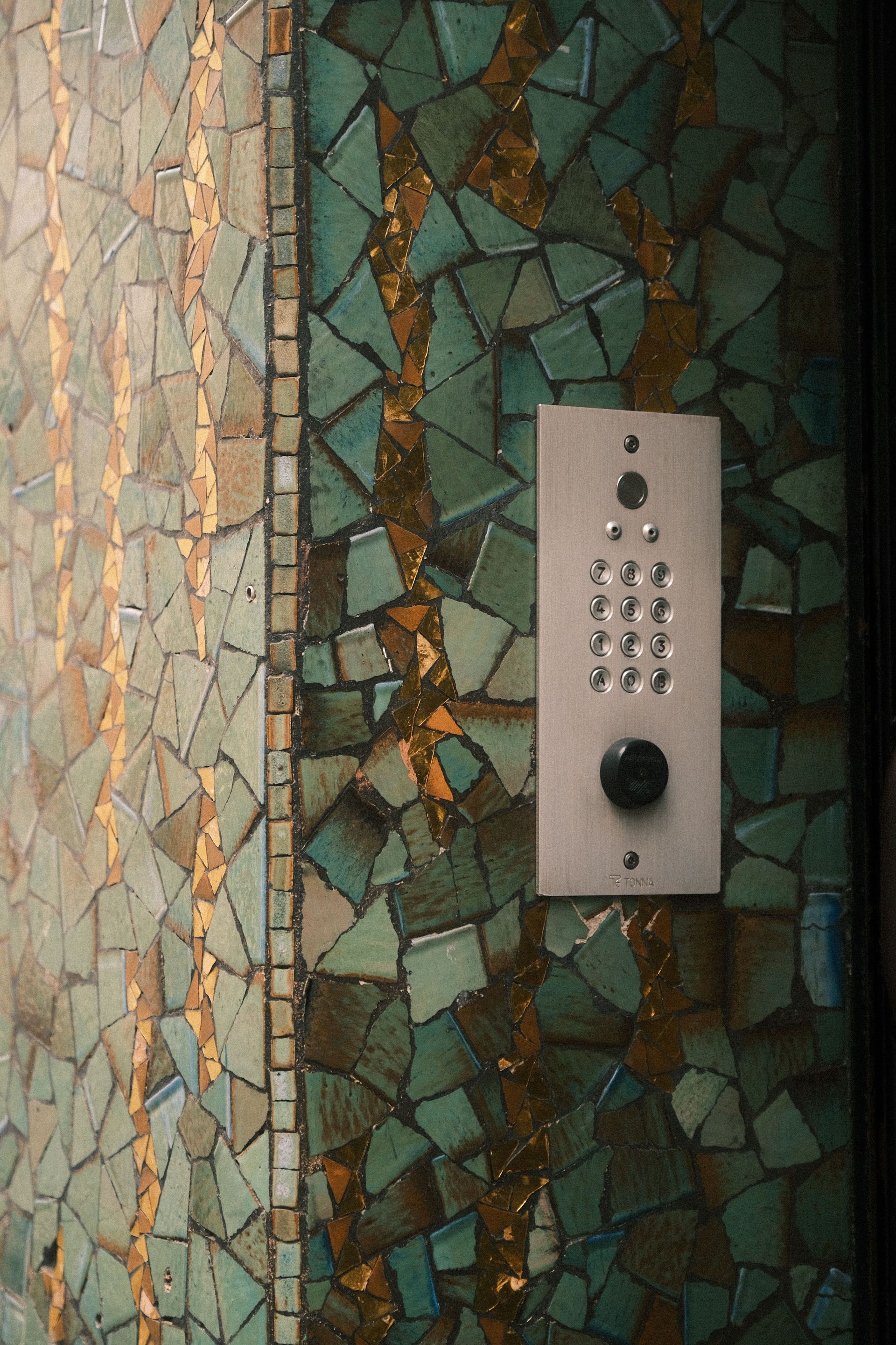 Pannello del citofono su una parete rivestita di mosaico con tastiera e pulsante nero.