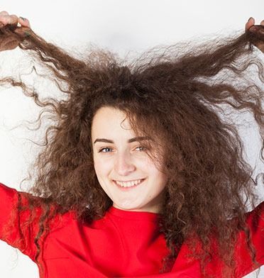 A person with long, voluminous, frizzy brown hair wears a bright red top and smiles while holding up strands of hair.