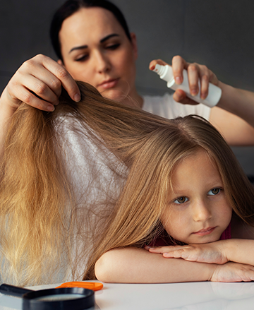 A person sprays a treatment into a young child’s hair while a magnifying glass sits on the table nearby.