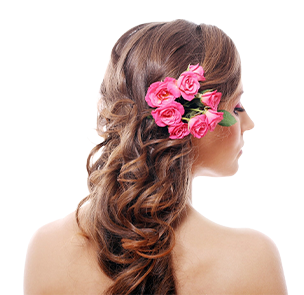 A person with long, wavy brown hair styled half-up and adorned with a cluster of bright pink roses.