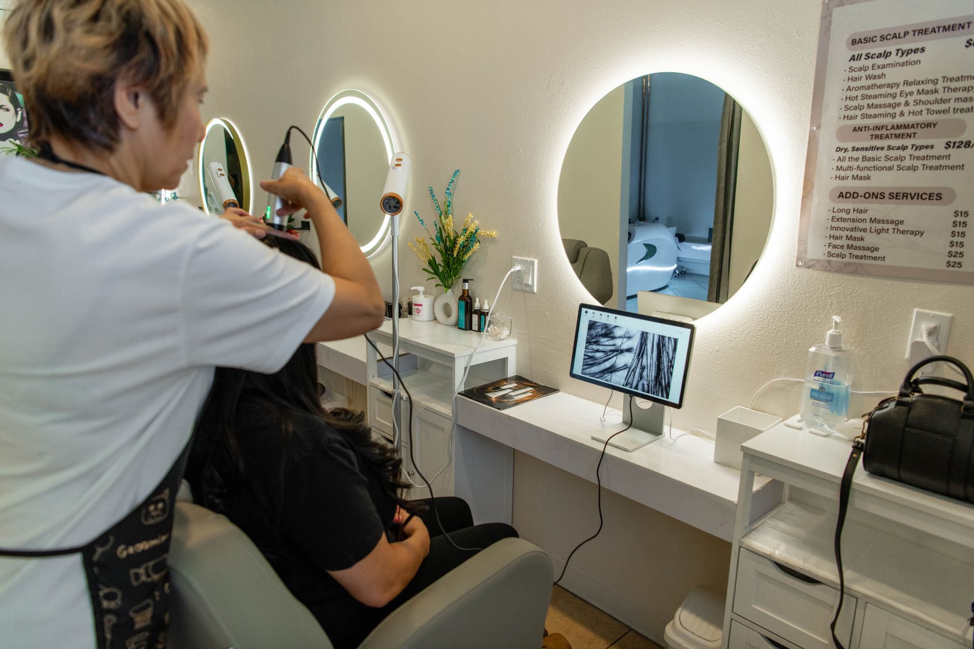 A hairstylist styles a client's hair in front of a mirror at a salon with a screen displaying hair imagery.