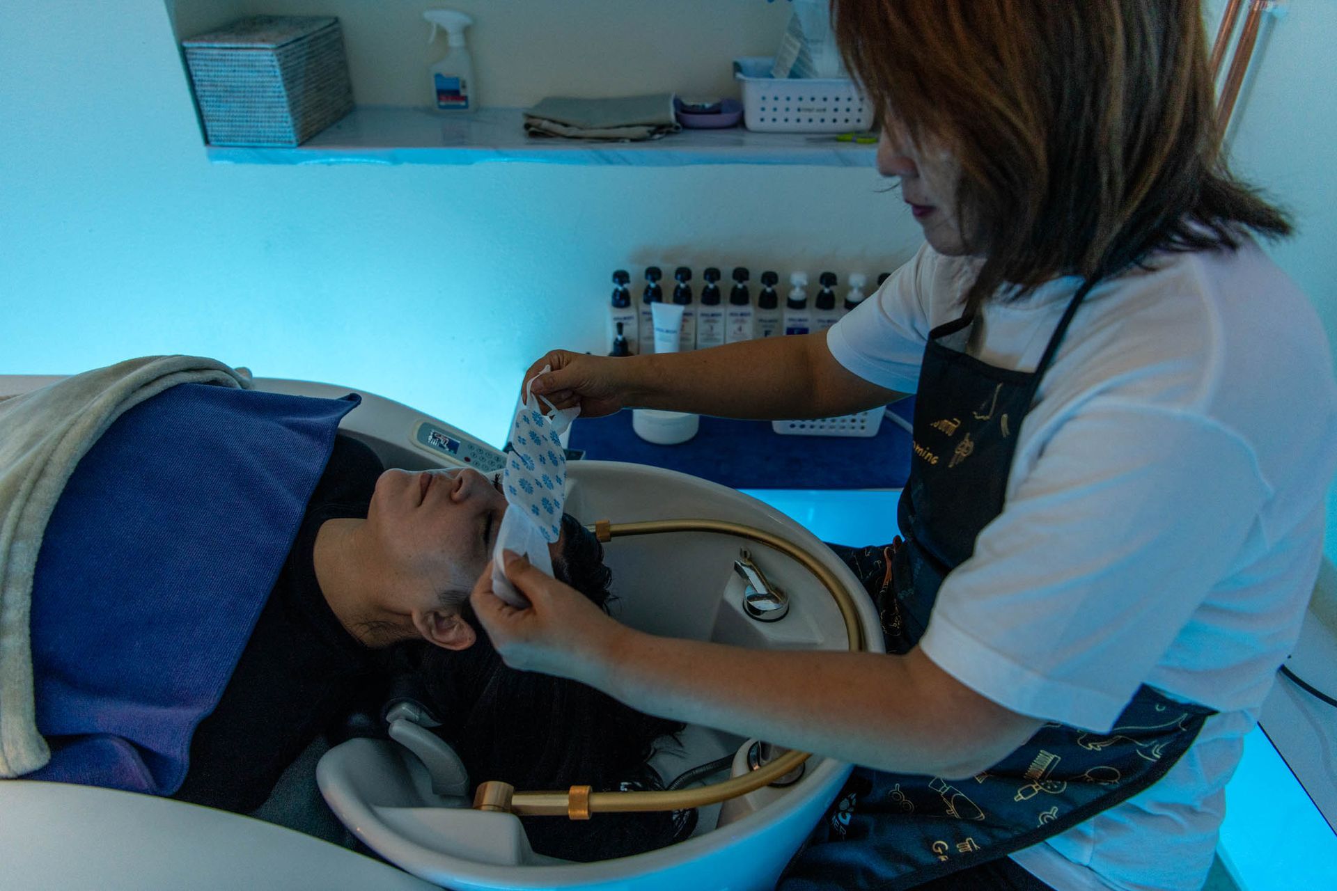 A stylist places a white face shield over a client's eyes while they recline at a shampoo bowl in a salon with blue lighting.