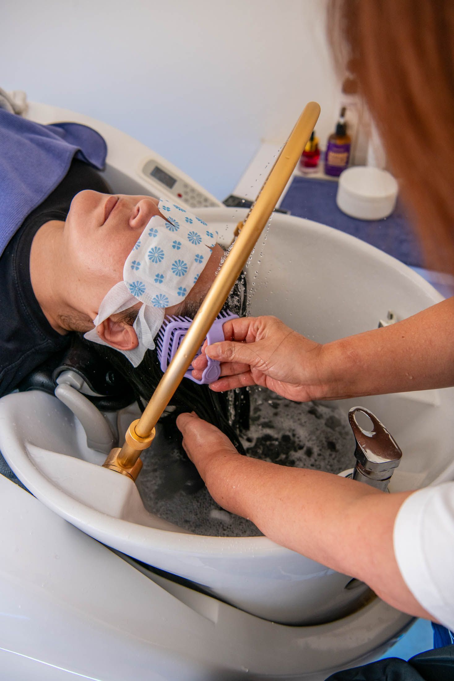 A stylist washes a client's hair in a salon sink, using a specialized spray tool to rinse suds from their covered eyes.