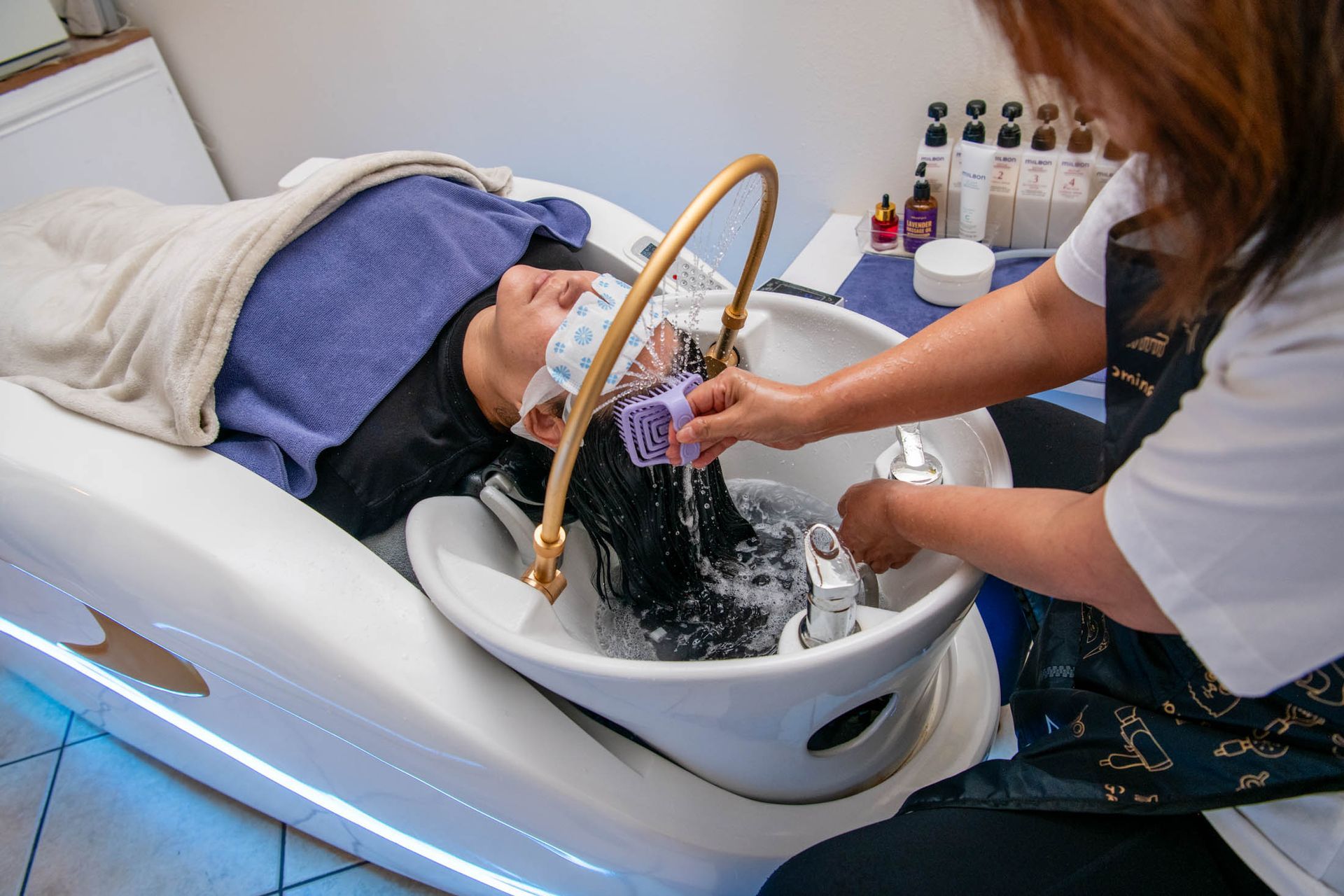 A stylist washes a client's hair in a salon sink, with water flowing from a curved metal arm over the hair.