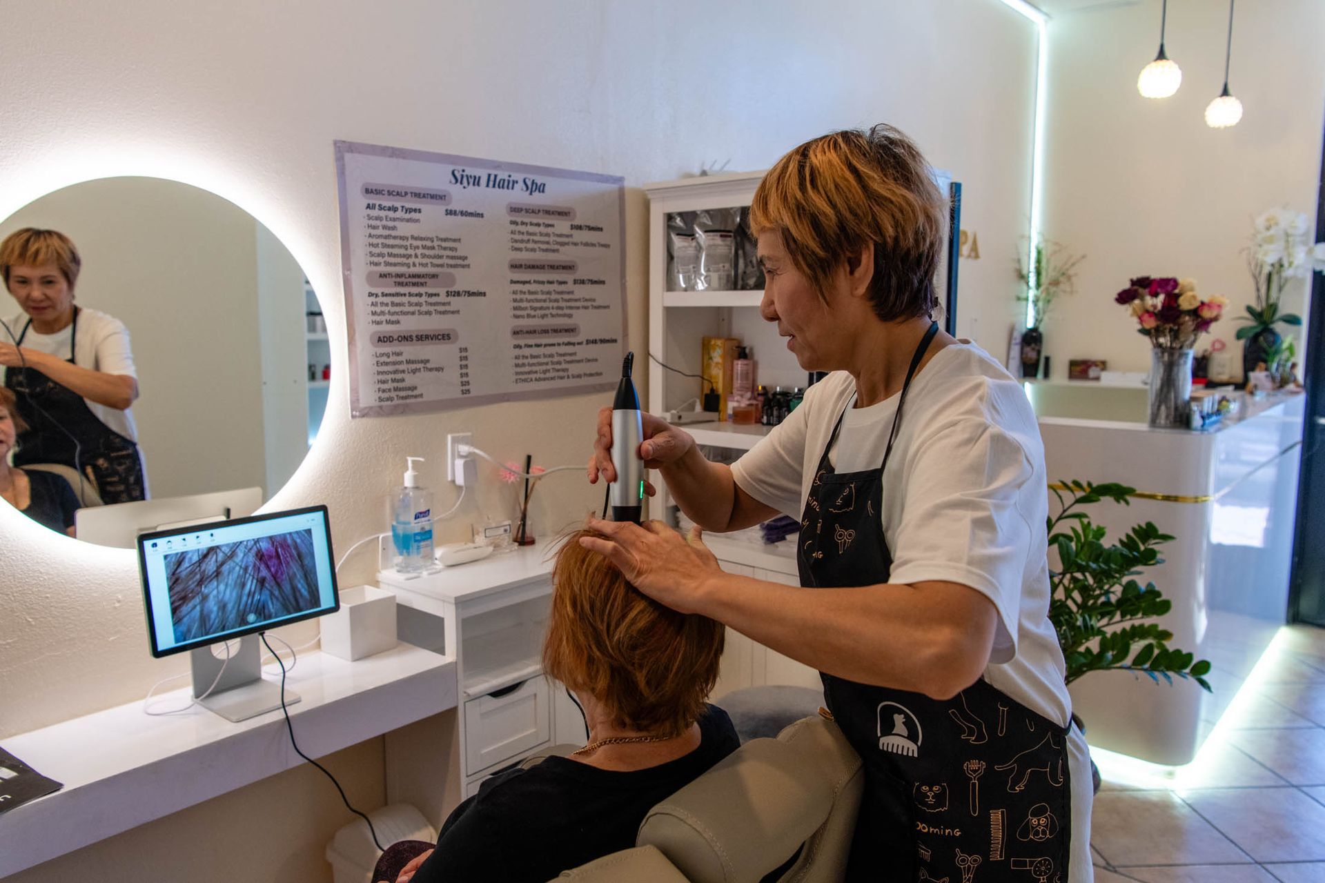A hair specialist uses a diagnostic device on a client's scalp, with the results displayed on a nearby monitor.