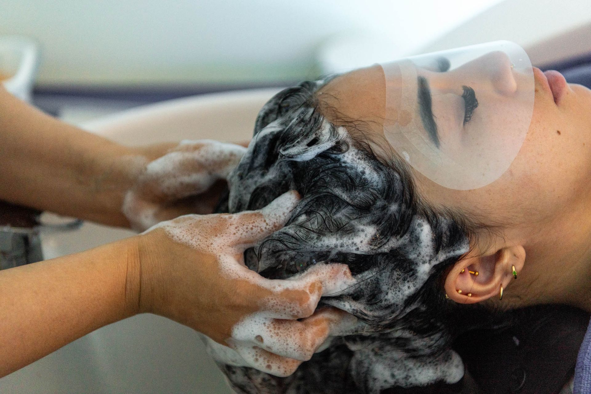 A person getting their hair washed in a salon sink while wearing a clear protective face shield.