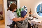 A massage therapist stands behind a seated person, gently massaging their neck in a bright spa or salon setting.
