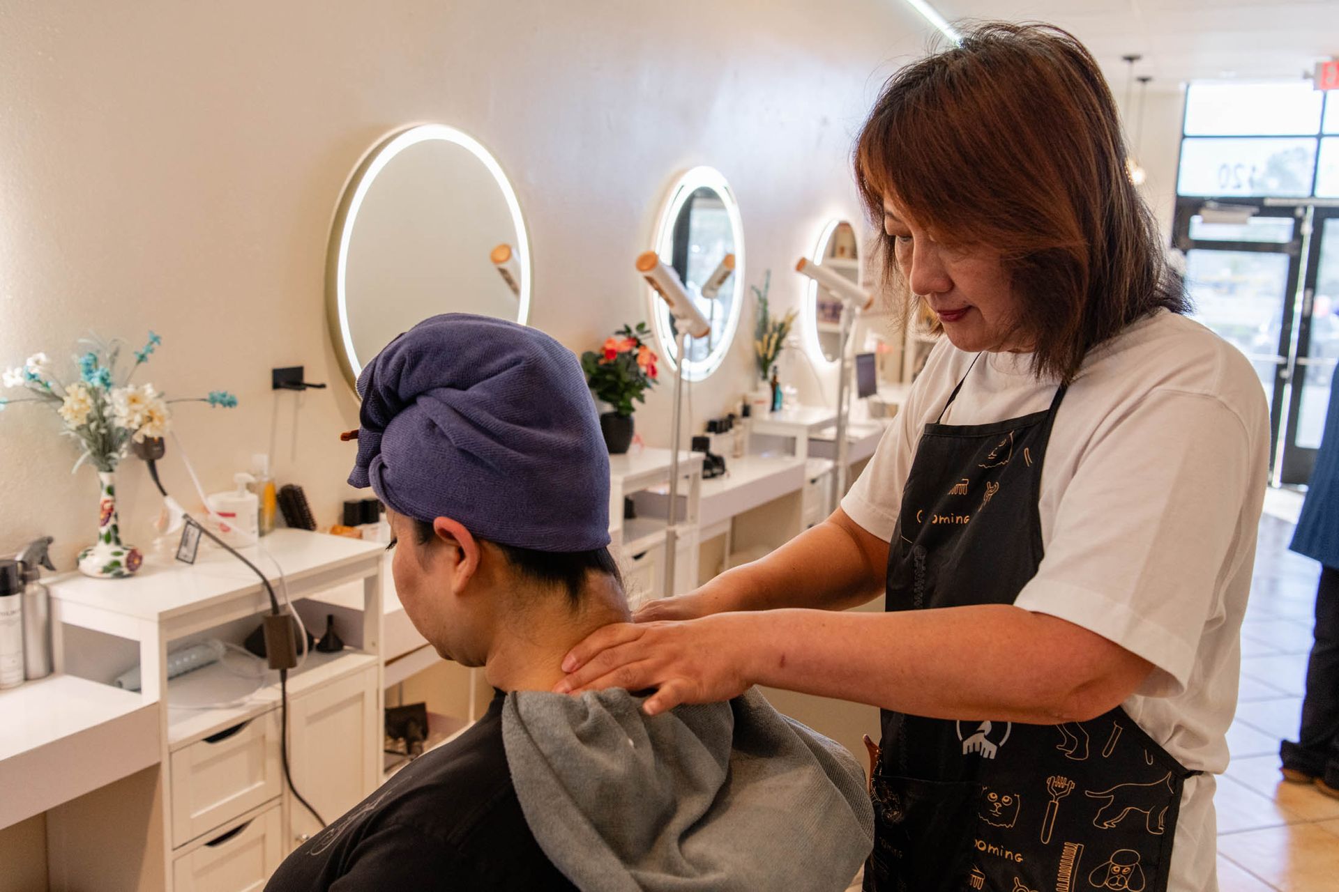 A stylist in a black apron massages the neck of a client wearing a purple hair wrap in a bright, modern salon.