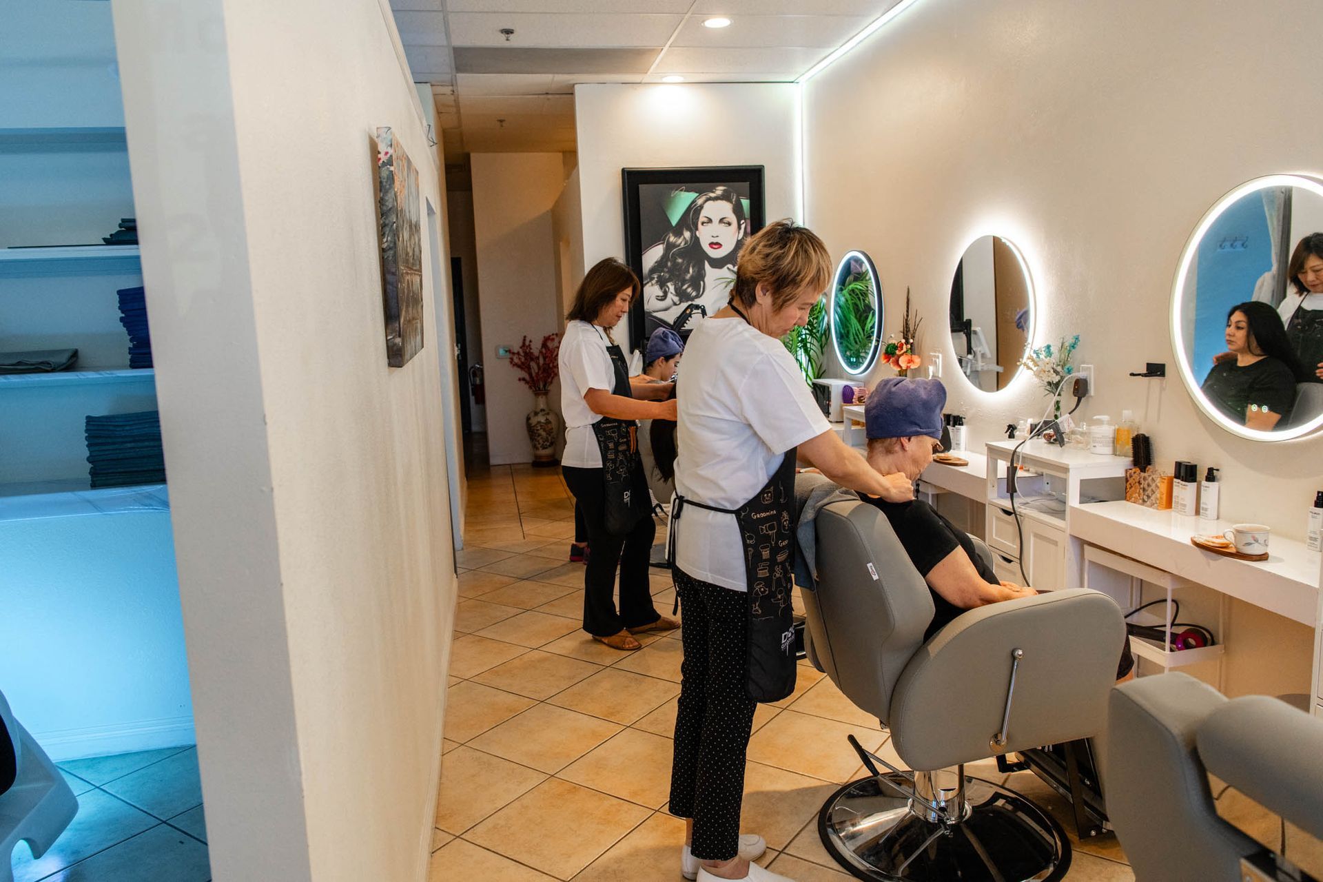 A hair salon interior with two stylists working on clients in chairs in front of lighted circular mirrors.