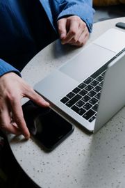 A person in a blue shirt sits at a round table, working on a silver laptop while holding a black smartphone.