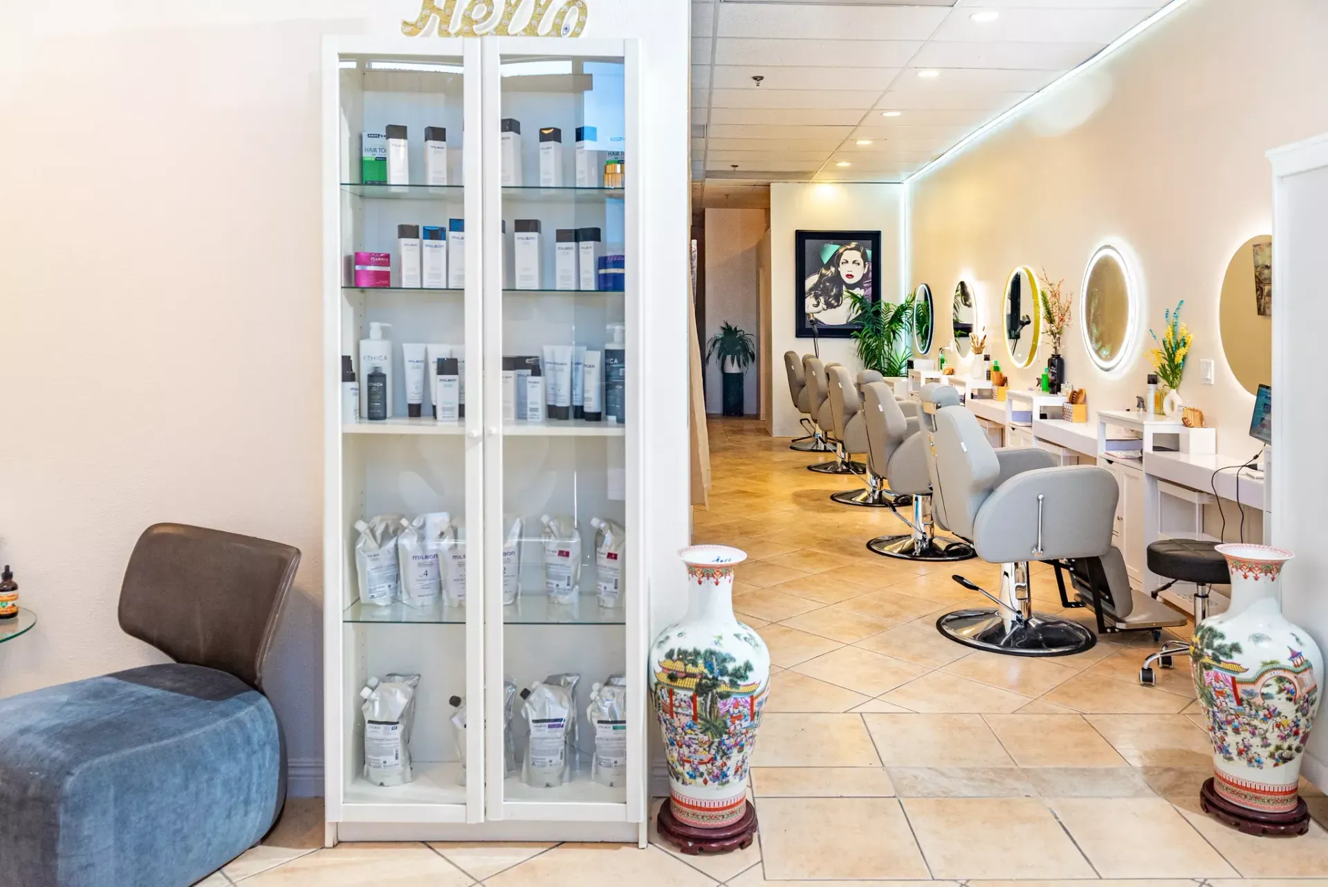A hair salon with a product display cabinet, two decorative floor vases, and a row of styling chairs with circular mirrors.
