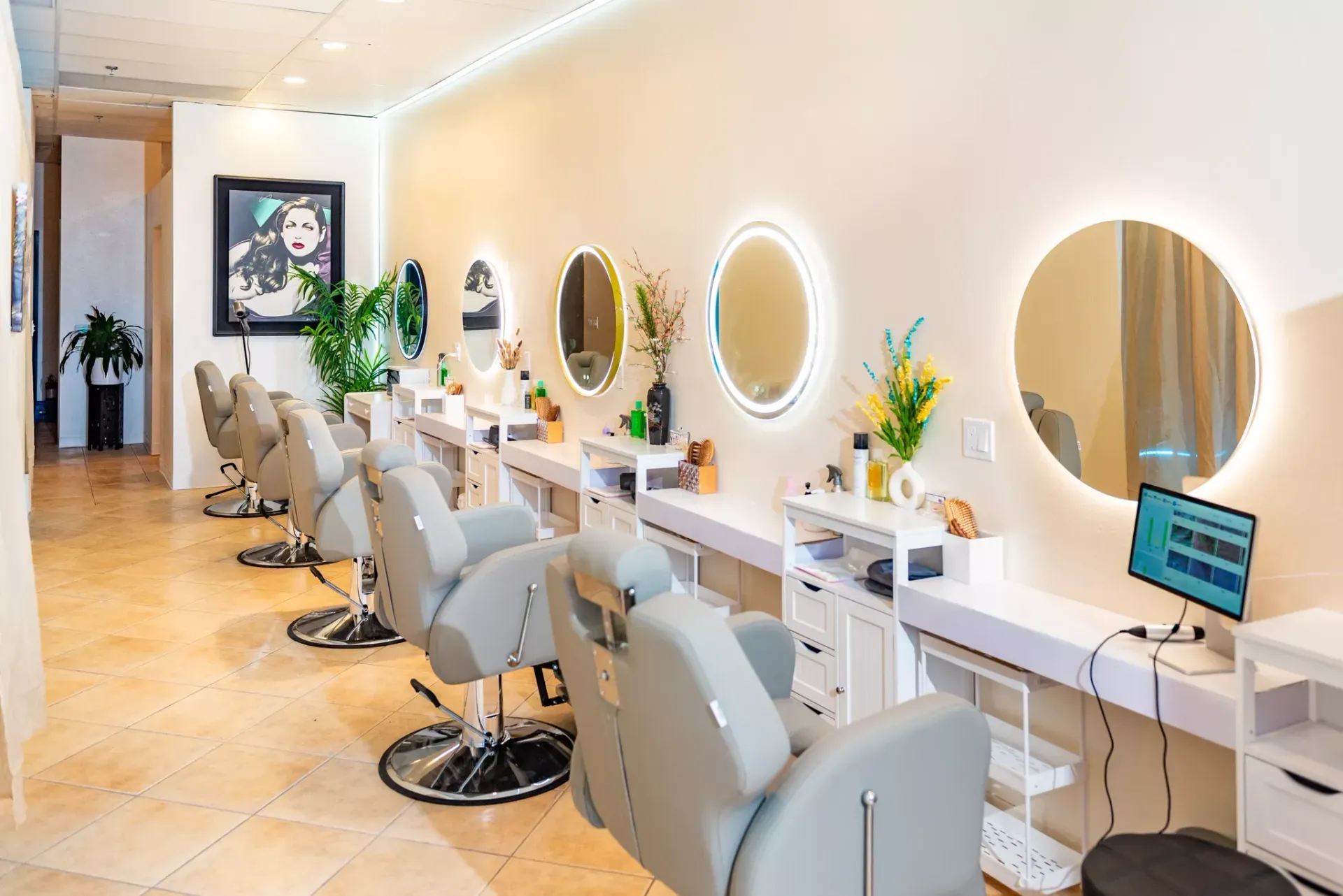 A bright salon with several grey chairs facing round, illuminated mirrors arranged along a white wall.