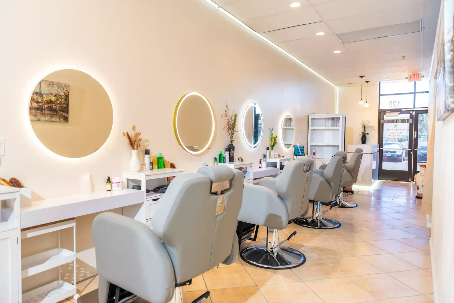 Interior of a bright, modern hair salon featuring light-colored styling chairs, circular backlit mirrors, and tile floors.