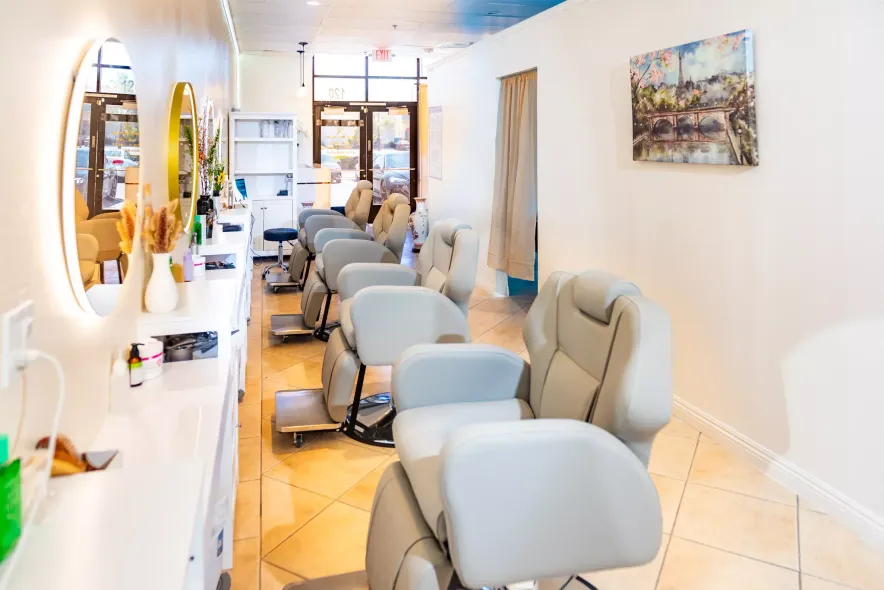 A bright salon interior features a row of light grey chairs, minimalist white counters, circular mirrors, and wall art.