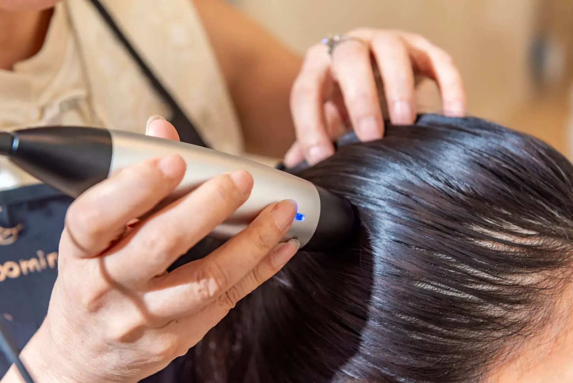 A hand uses a specialized handheld device to apply treatment to a person's scalp and dark hair.