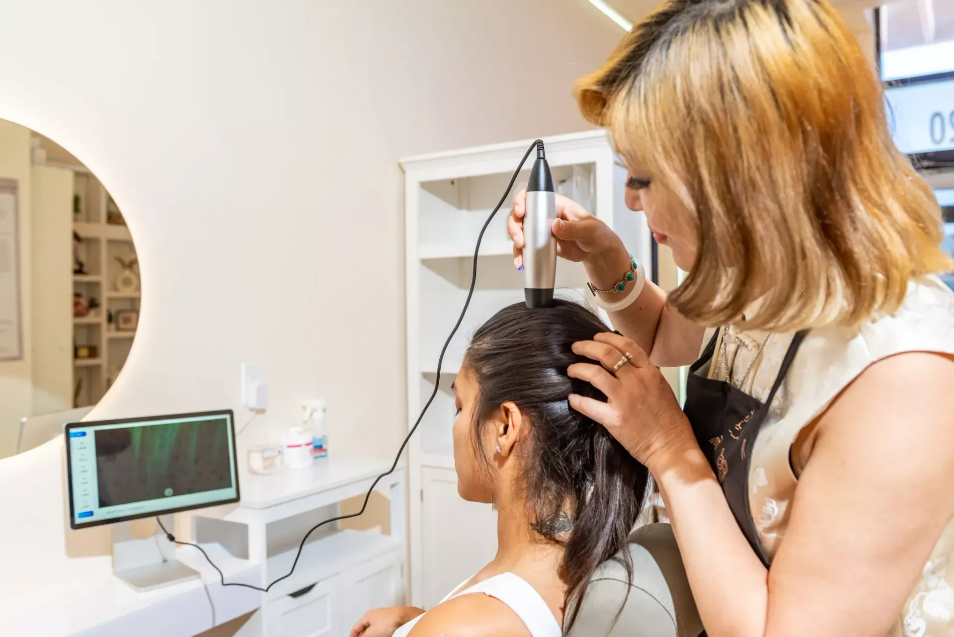 A professional uses a scalp analysis device on a client, with the results displayed on a monitor in a bright salon.