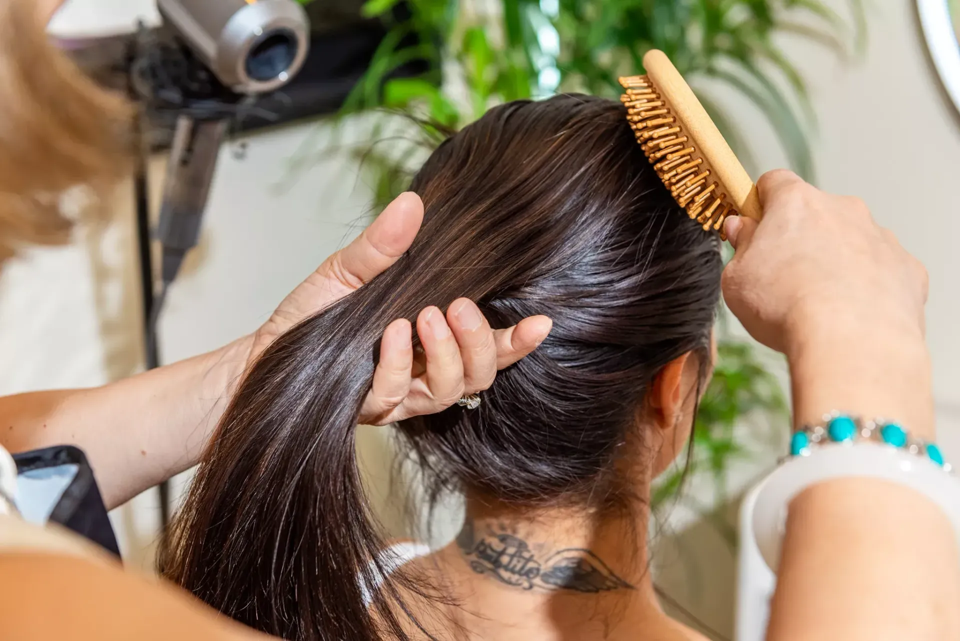 A person with a neck tattoo holds a hairbrush while their hair is styled and brushed from behind.