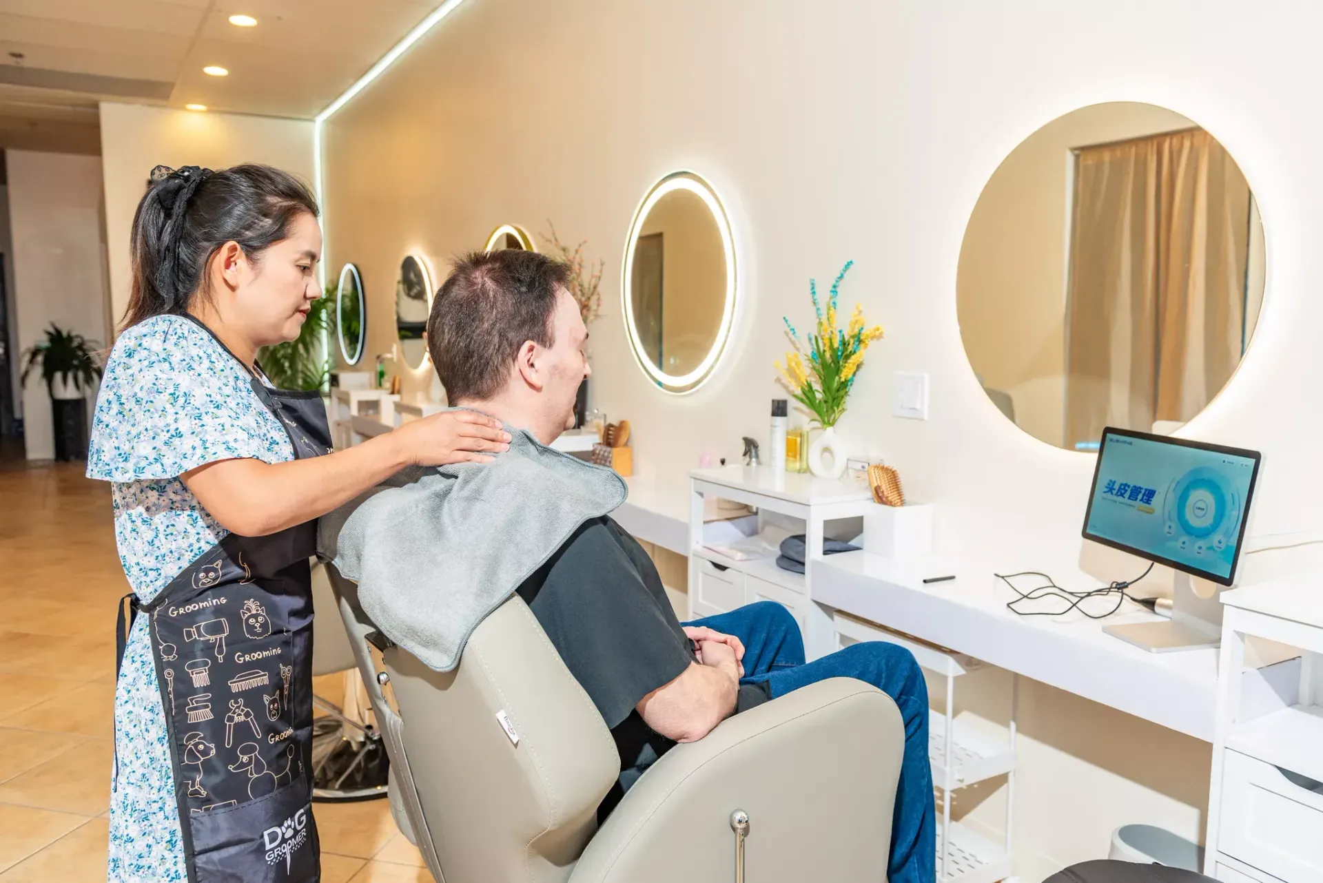 A stylist in a patterned apron adjusts a gray drape around a client sitting in a salon chair before a circular mirror.