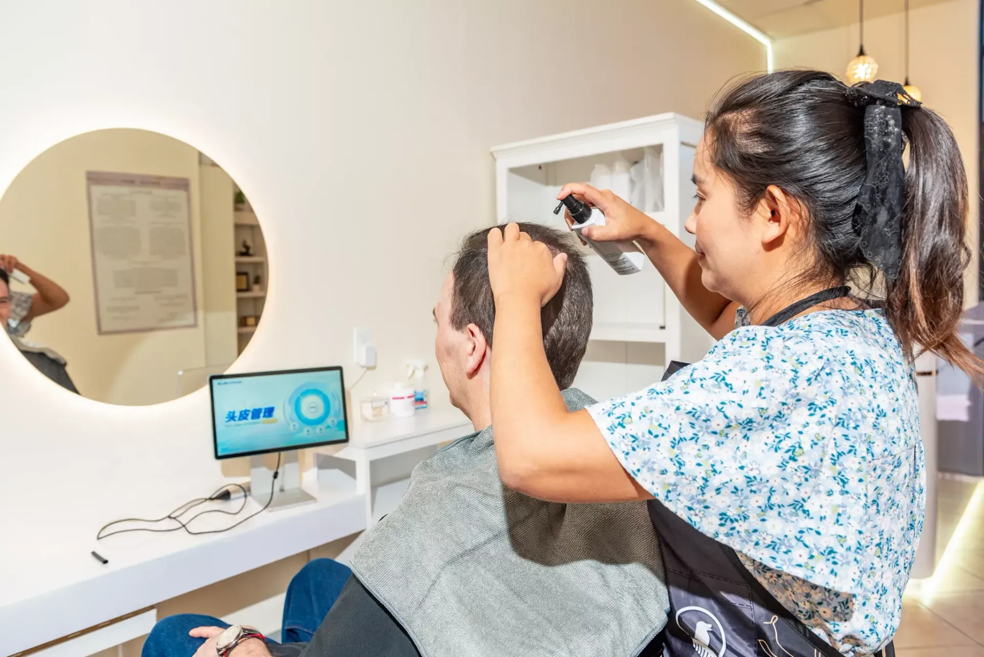 A hair specialist applies treatment to a client's scalp in a clinic with a screen displaying hair growth data.