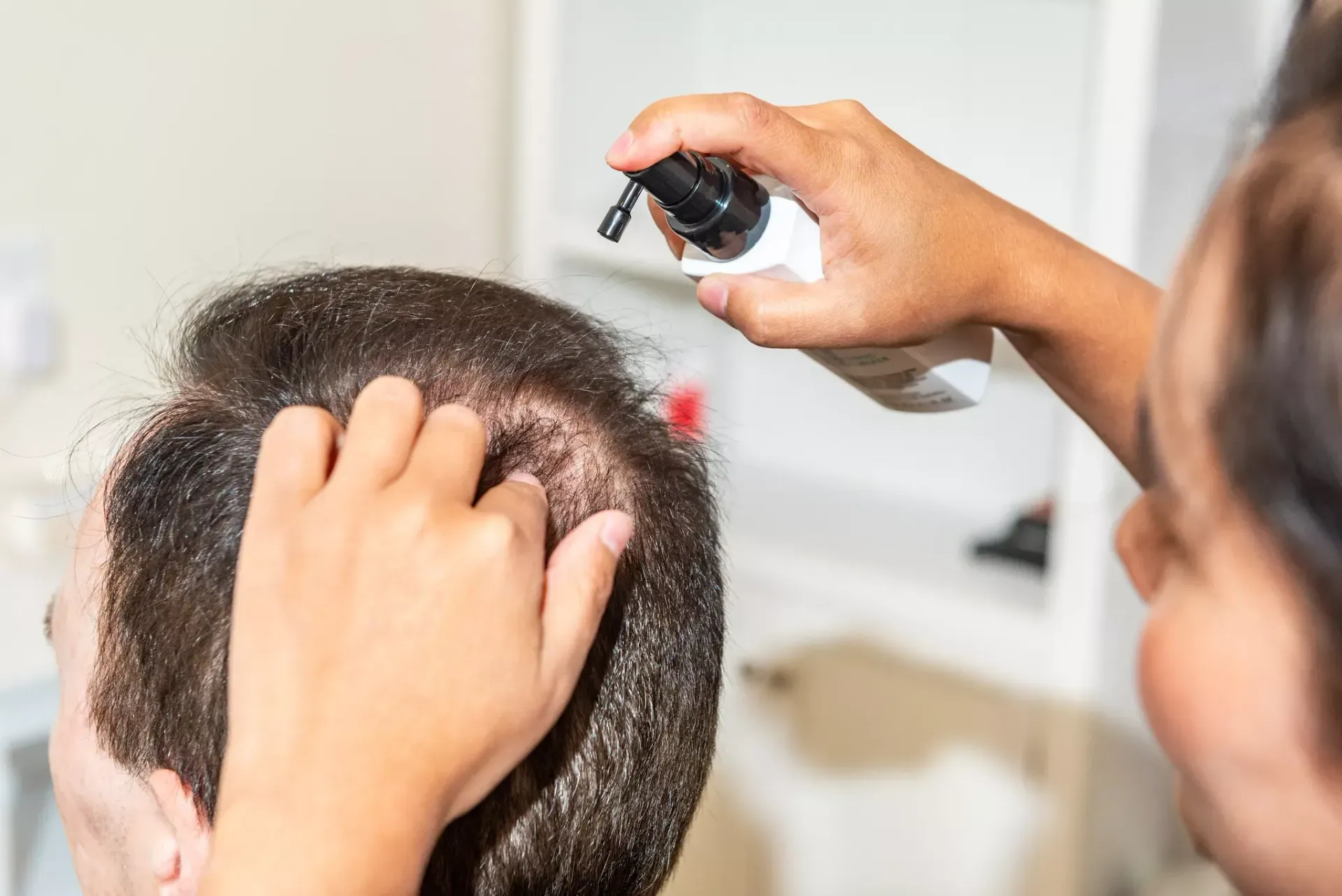 A person applies spray treatment to the scalp of another person, using their fingers to massage the area.