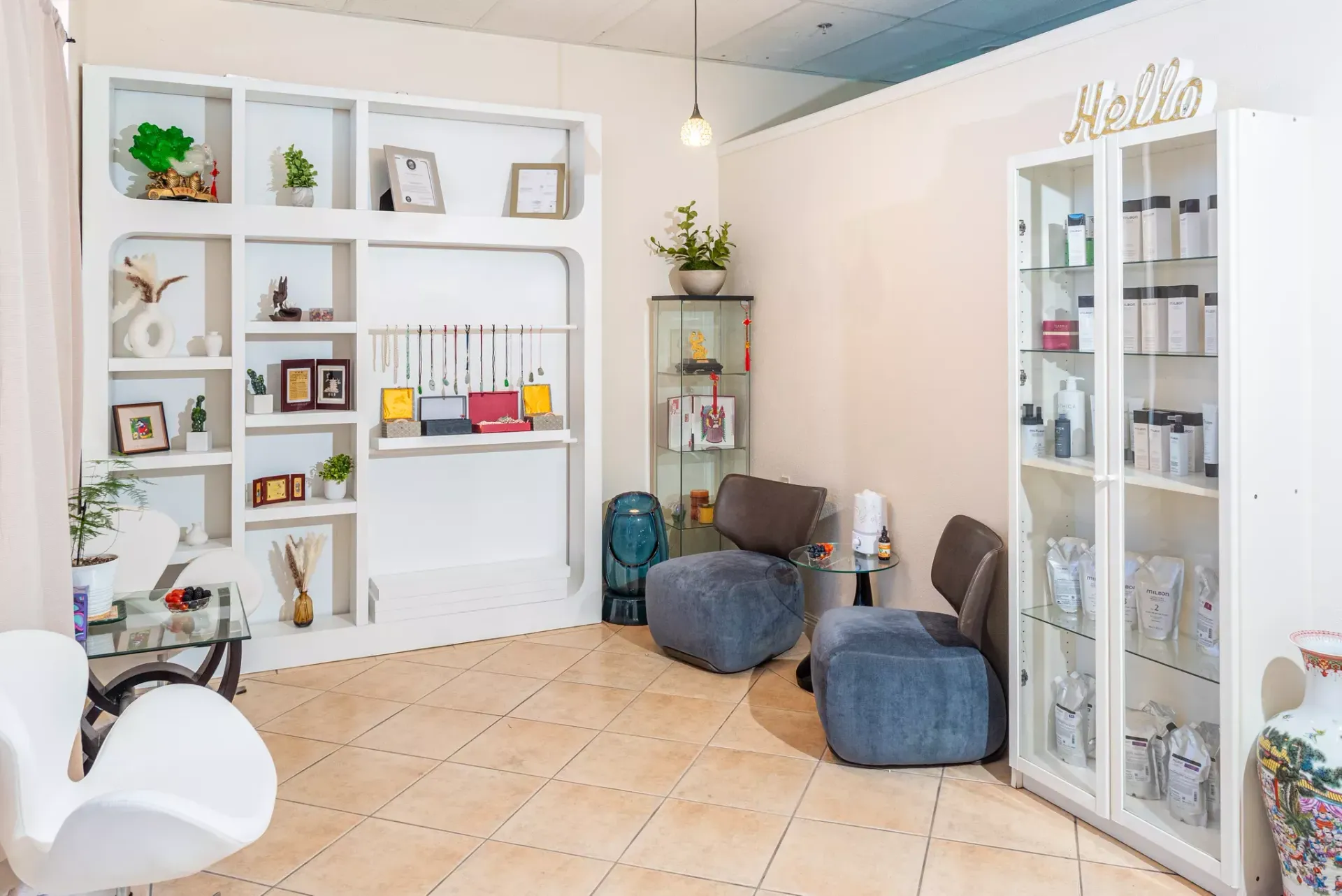 A bright boutique retail space with white shelving, a glass display cabinet, two grey armchairs, and light tiled floors.