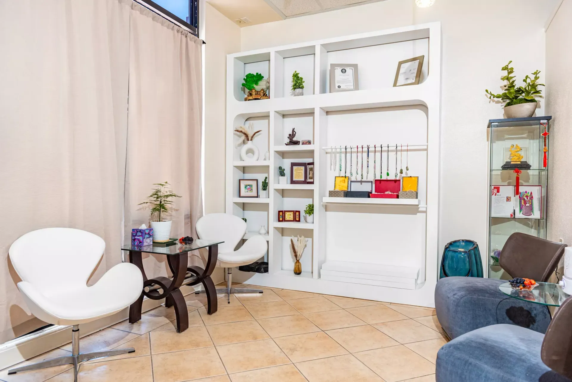 Modern waiting area with white swan-style chairs, a shelving unit, a glass display case, and beige tiled floors.
