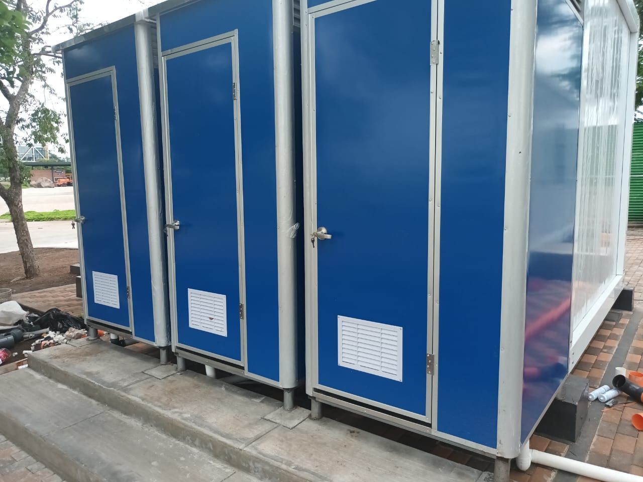 Tres baños portátiles azules con rejillas de ventilación y puertas blancas. Están colocados sobre una base de hormigón.
