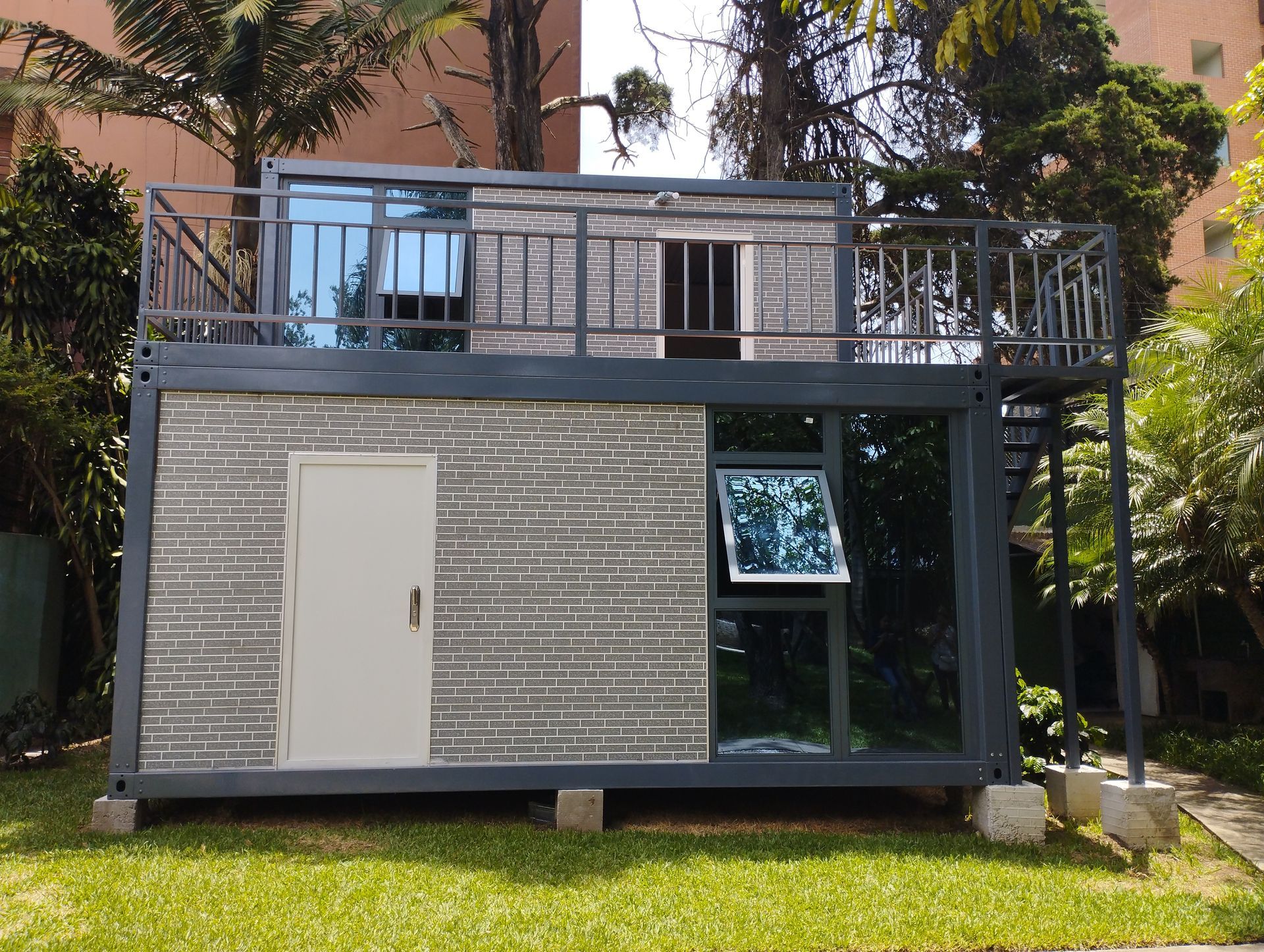 Edificio gris de dos plantas construido con contenedores marítimos, con ventanas y balcón, situado sobre un césped.