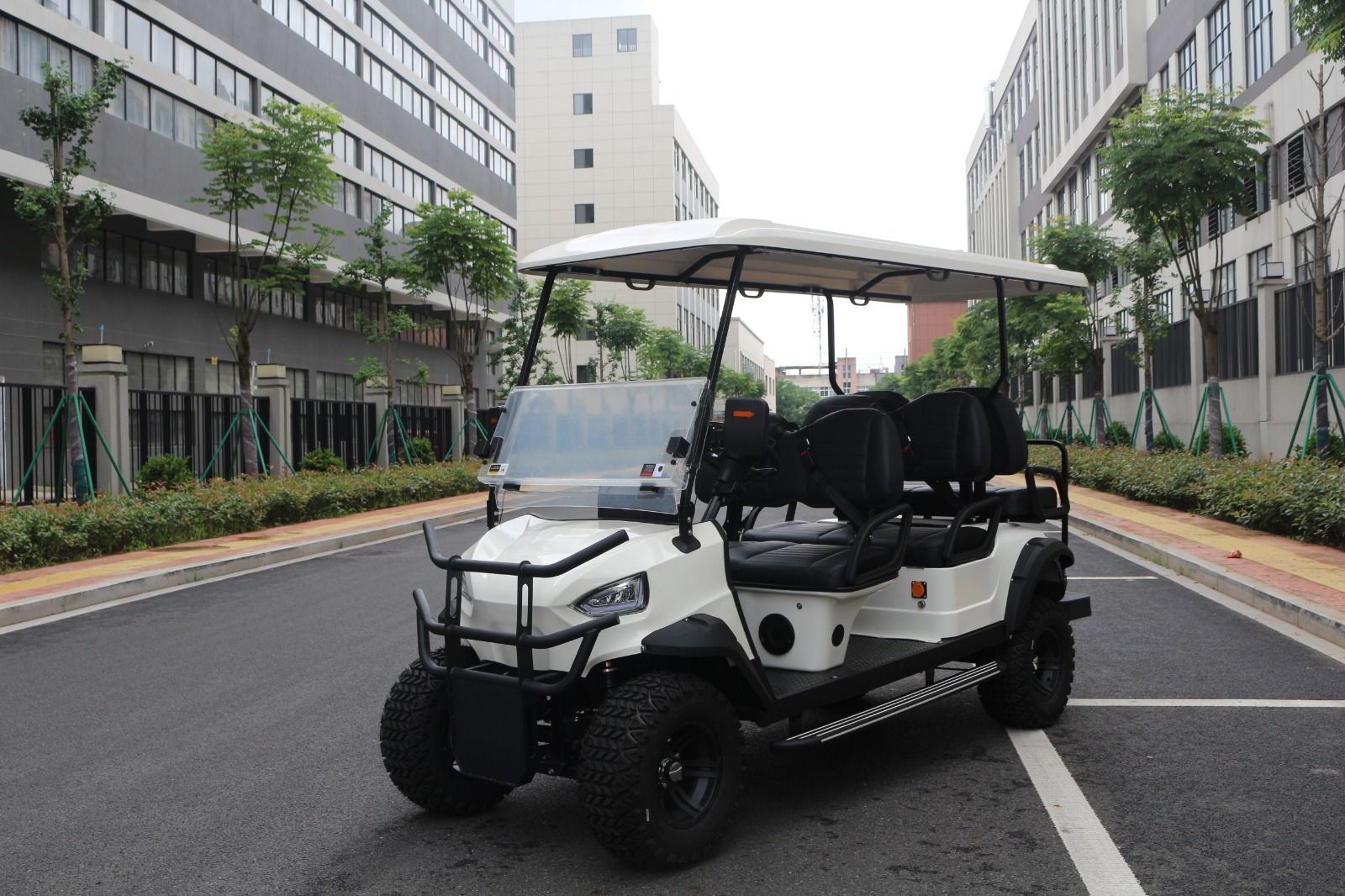Carrito de golf blanco con neumáticos negros estacionado en una carretera asfaltada. Edificios y árboles al fondo.