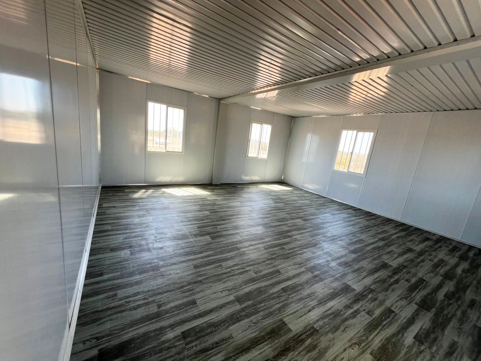 Habitación vacía con suelo de imitación madera, paredes blancas, tres ventanas y techo blanco.