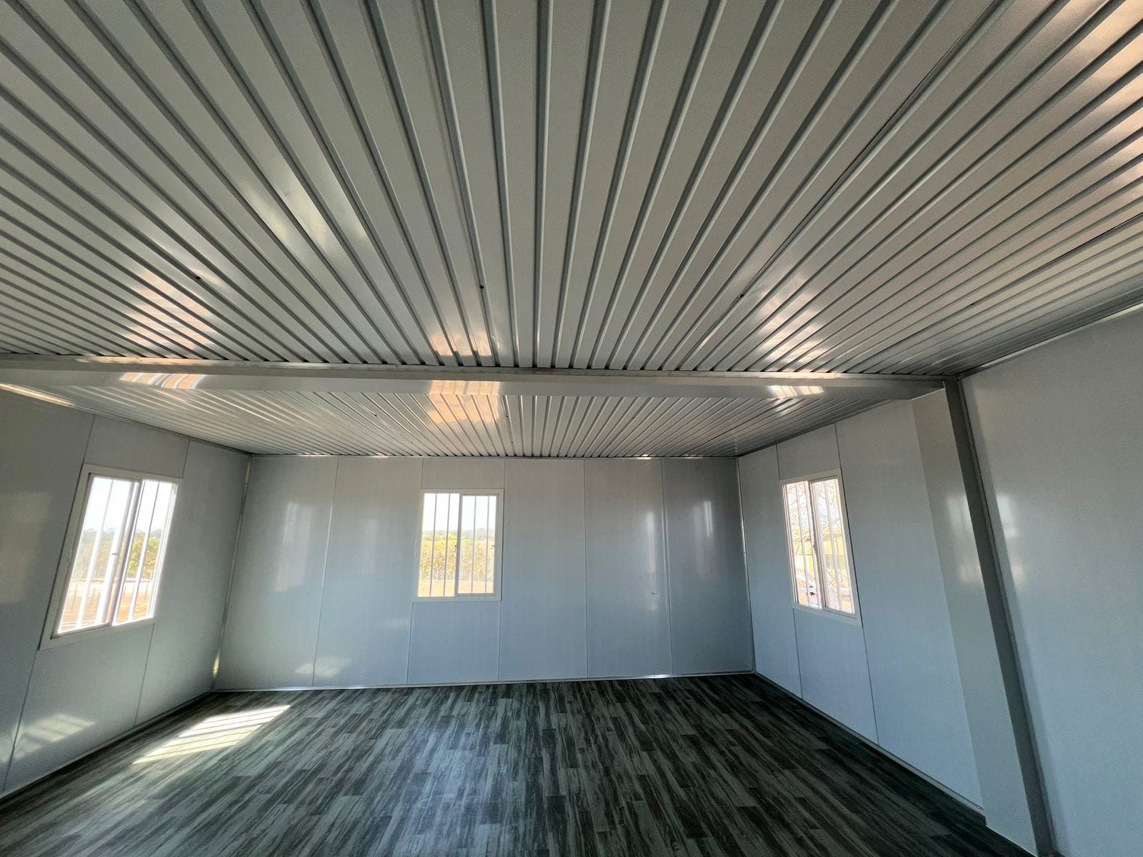 Interior de un edificio prefabricado blanco con tres ventanas, suelo con aspecto de madera y techo ondulado.