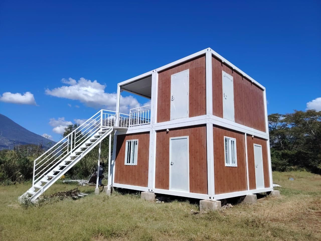 Edificio modular de dos plantas con escaleras en un campo de hierba, con cielo despejado al fondo.