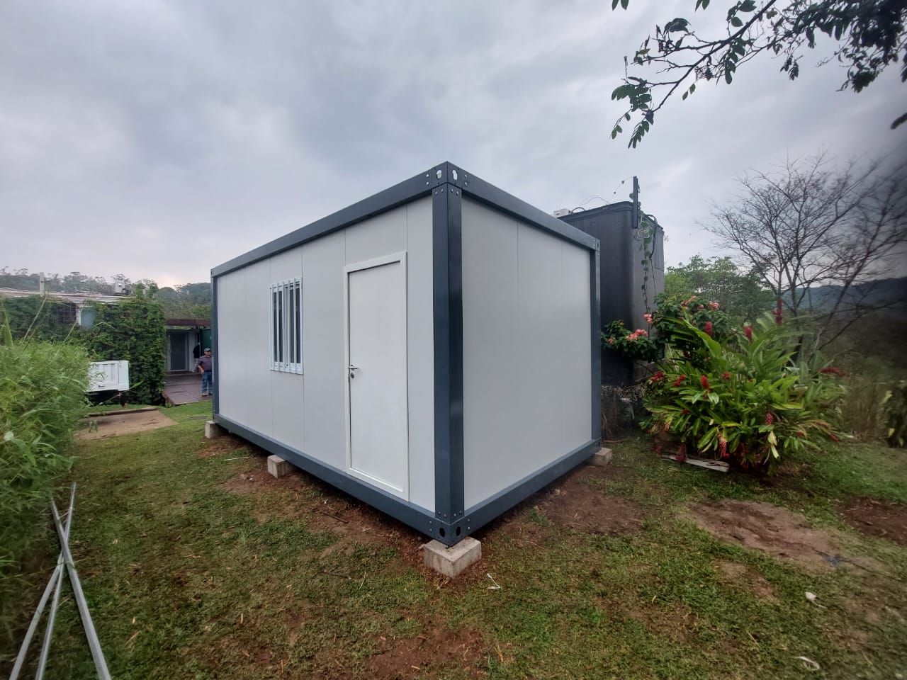 Edificio modular gris con puerta y ventana, situado sobre bloques de hormigón al aire libre