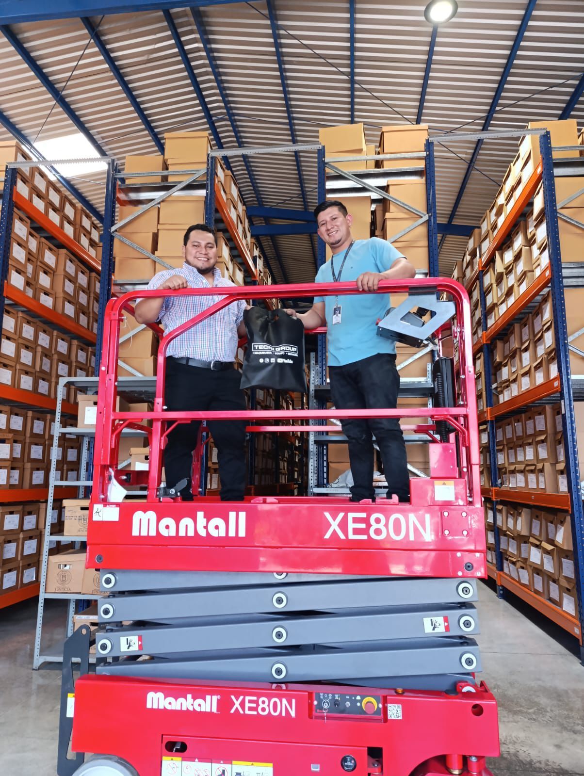 Dos hombres en una plataforma elevadora Mantall XE80N roja en un almacén